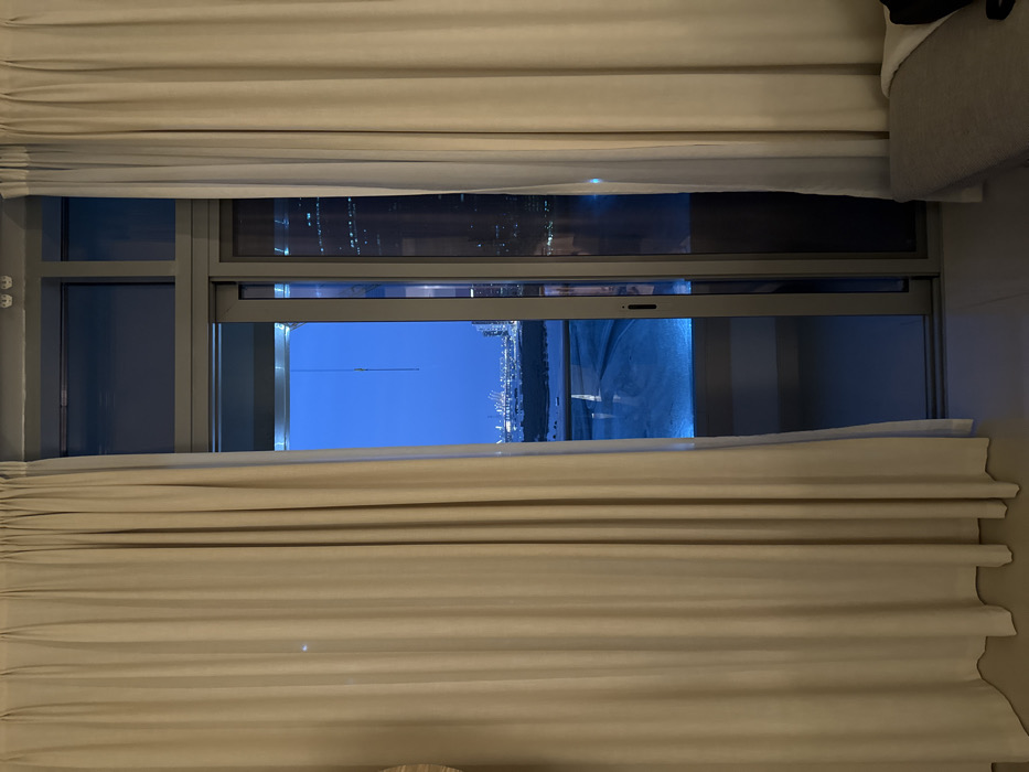 The image captures an interior perspective looking outward through a large, multi-paneled glass door and fixed window at twilight or night in Abu Dhabi, United Arab Emirates. Beige, pleated curtains are partially drawn open, framing the central glass portal.
Visible through the glass is an exterior balcony featuring a luminous, rectangular blue-lit pool occupying the lower-middle frame. Beyond the balcony, a dark landmass or shoreline is present, leading to a distant cityscape marked by numerous artificial lights and tall structures. A prominent port or industrial zone, identified by multiple illuminated cranes, is discernible on the horizon. The sky above is a uniform deep blue. A slender, light-colored vertical wire or cable descends from the upper glass frame into the exterior view.
The indoor environment includes a segment of a light-grey upholstered bench with a dark cushion in the lower right foreground and a circular, woven light-colored object partially visible on the left edge. The interior floor appears light-colored and unadorned. No human figures or discernible actions are depicted.