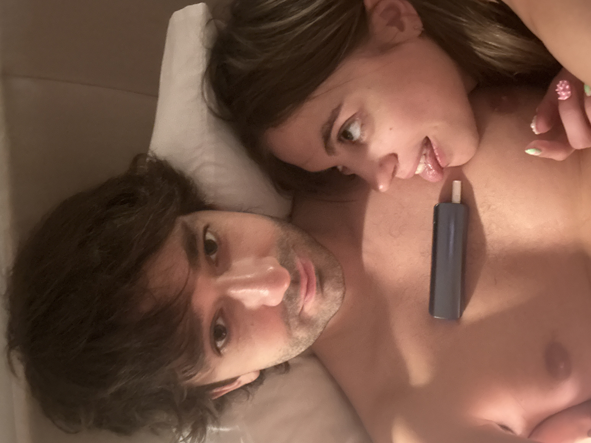 The image is a close-up selfie of a man and a woman lying in bed. The man is in the foreground, looking at the camera with messy dark hair and stubble. A small dark electronic device is resting on his bare chest. To the right of the man, the woman is visible from the side, looking upwards and sticking her tongue out. Her hair is brown and long, and she is wearing a ring on her finger with light green nail polish. The background appears to be a pillow and bedding. There is no information in the image to suggest a location in Moscow, Russia.