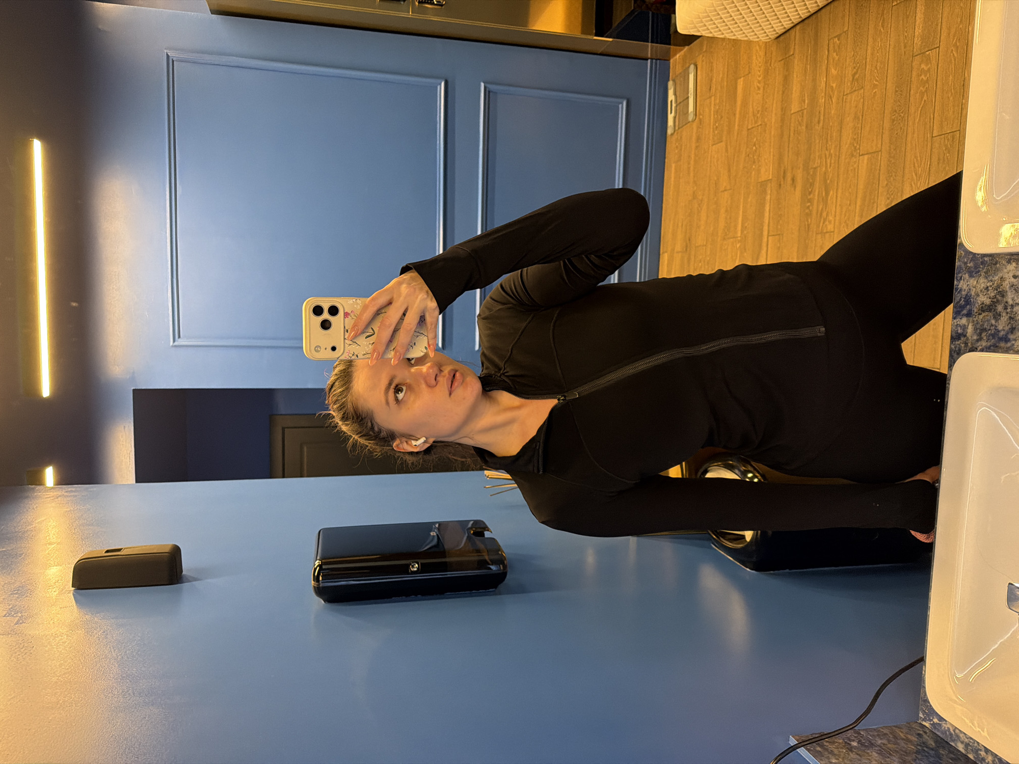 A woman in black leggings and a black zip-up top is taking a selfie with a smartphone in a bathroom. She has blonde hair pulled back, and an AirPod is visible in her ear. The bathroom features a blue vanity with a dark brown dispenser and a shiny black container on the counter. The background includes a blue wall with a rectangular recessed panel design and a horizontal light fixture. The floor is wood. The location is likely a bathroom in a building in Belgrade, Serbia.