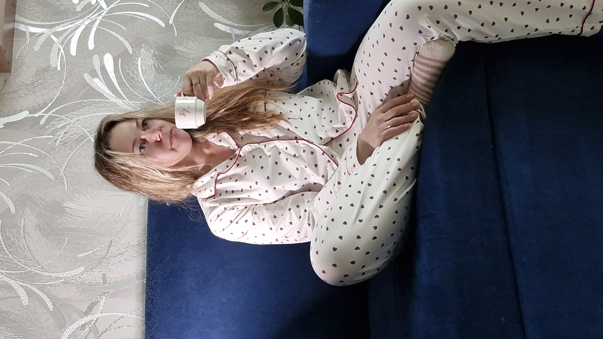 A woman is reclining on a blue sofa, dressed in pajamas patterned with small brown hearts, and striped socks. She is holding a white mug. The background includes a wall with an ornate silver design and a glimpse of a plant. The exact location is not discernible from the image.