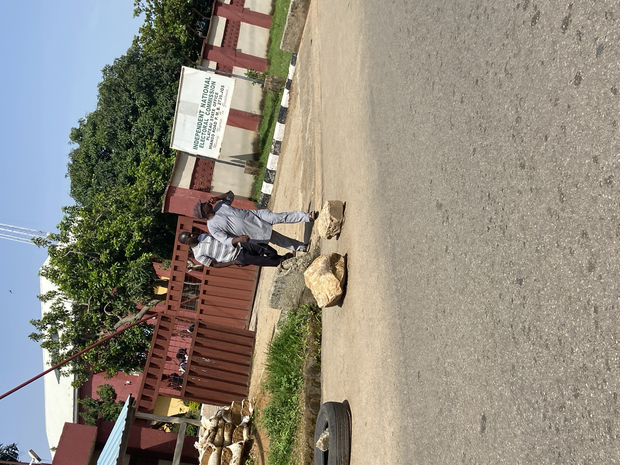 The image depicts an outdoor scene on a paved road in Jos, Nigeria, in front of a building identified by a sign as the "INDEPENDENT NATIONAL ELECTORAL COMMISSION PLATEAU STATE OFFICE MIANGO ROAD P. M. B. 2739, JOS Moving Nigeria to Greatness". Two men are walking on the road. One man, on the left, is wearing a grey traditional outfit and a cap, and is holding his hand to his mouth, possibly speaking on a phone. The other man, on the right, is wearing a striped t-shirt and dark trousers. In the foreground of the road, there are two large rocks and a deflated tire. Behind the men, there is a brown metal gate leading into the compound, and further back, trees and a red building with a white roof are visible. There are also piles of sandbags to the left of the gate. The sky is clear and blue.