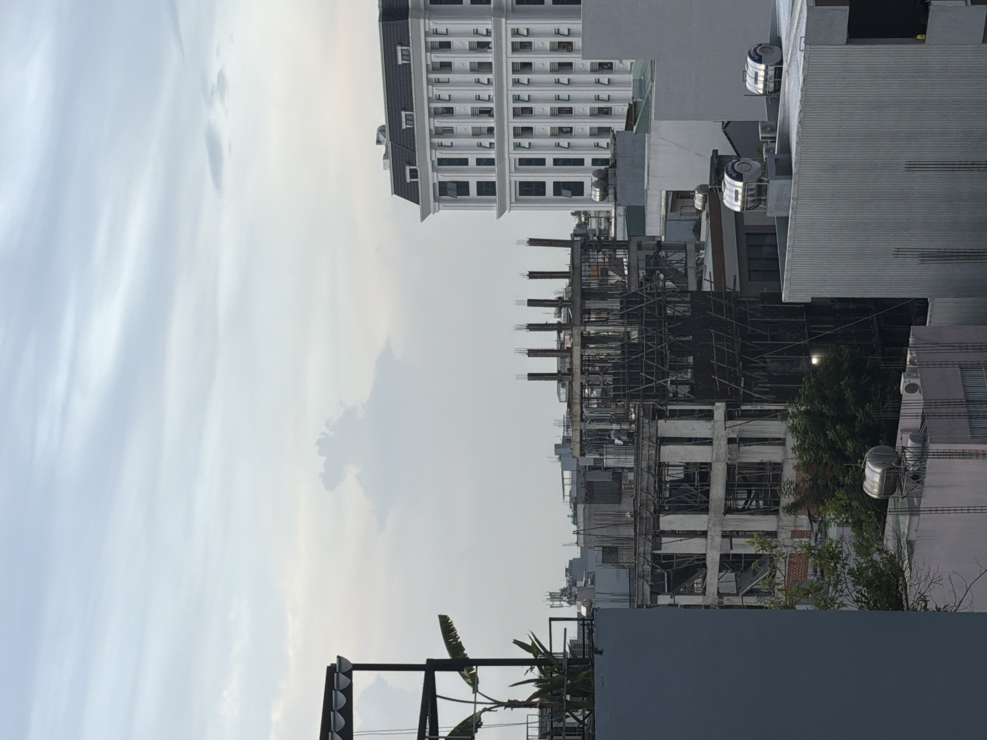 This image captures a cityscape in Đà Nẵng, Vietnam, characterized by overcast skies and the presence of multiple buildings in various stages of construction and completion. In the foreground, a portion of a blue-gray building occupies the left side of the frame, with a dark metal structure and a banana plant on its rooftop. To the right, a partially constructed concrete building is visible, featuring exposed rebar and scaffolding, with one person standing near the top. Further into the background, a pristine white building with a dark, gabled roof stands prominently, adorned with numerous windows. Several other buildings, some appearing to be residential or commercial, are scattered throughout the scene, with some featuring rooftop water tanks and air conditioning units. The overall impression is one of ongoing urban development against a muted sky.