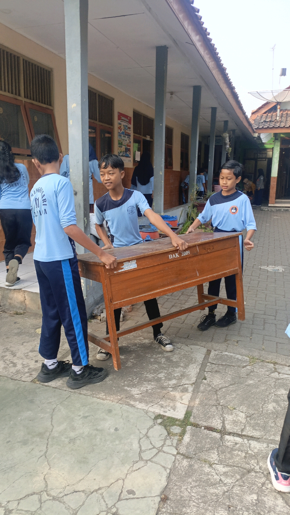 In Salatiga, Indonesia, two male students in light blue school uniforms are carrying a wooden school desk. One student, on the left, wears a blue long-sleeved shirt with "SD NGLUWARGA 01" visible on the back and dark blue pants with a light blue stripe. He is barefoot. The other student, on the right, wears a light blue short-sleeved shirt with a dark blue collar and dark blue pants. Both students are gripping the desk, positioned at the front corners, and appear to be moving it outdoors on a paved walkway. The desk is made of wood and has "DAK 2009" printed on its front. Behind them, a covered walkway with pillars and classrooms with barred windows can be seen. The ground is cracked concrete. The sky is overcast. Several other people are visible in the background, including individuals in similar uniforms.