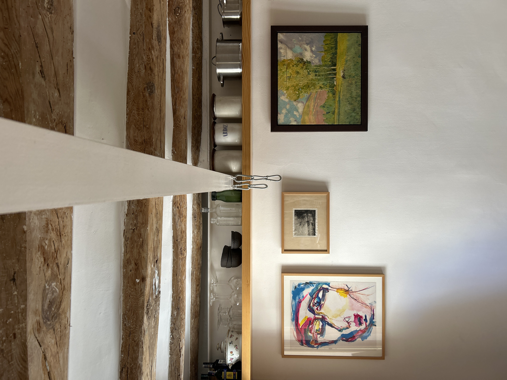 The image depicts an interior scene with a rustic aesthetic, likely a kitchen or dining area. Overhead, exposed wooden beams crisscross the ceiling, with white plaster filling the spaces between them. A white fabric element hangs down, suspended by silver carabiners.
Below the beams, a wooden shelf displays various kitchen items. These include bottles of what appear to be wine or spirits on the left, a lidded decorative bowl, several wine glasses, a stack of dark bowls, and glass bottles. Further to the right are three white canisters, one labeled "ARROZ" (rice in Spanish), and several stainless steel pots and pans.
The wall below the shelf is painted white and features three framed artworks. On the left, a colorful abstract portrait of a face is prominently displayed. To its right, a smaller, rectangular frame contains a black and white landscape photograph. On the far right, a larger framed oil painting depicts a pastoral scene with a field, trees, and a distant hillside under a cloudy sky.
The overall impression is of a well-lived-in space that combines functional kitchen elements with decorative art, suggesting a home with character and artistic taste. The presence of "ARROZ" on a canister might hint at a Spanish influence, but the image itself does not provide specific geographical markers for Barcelona, Spain.