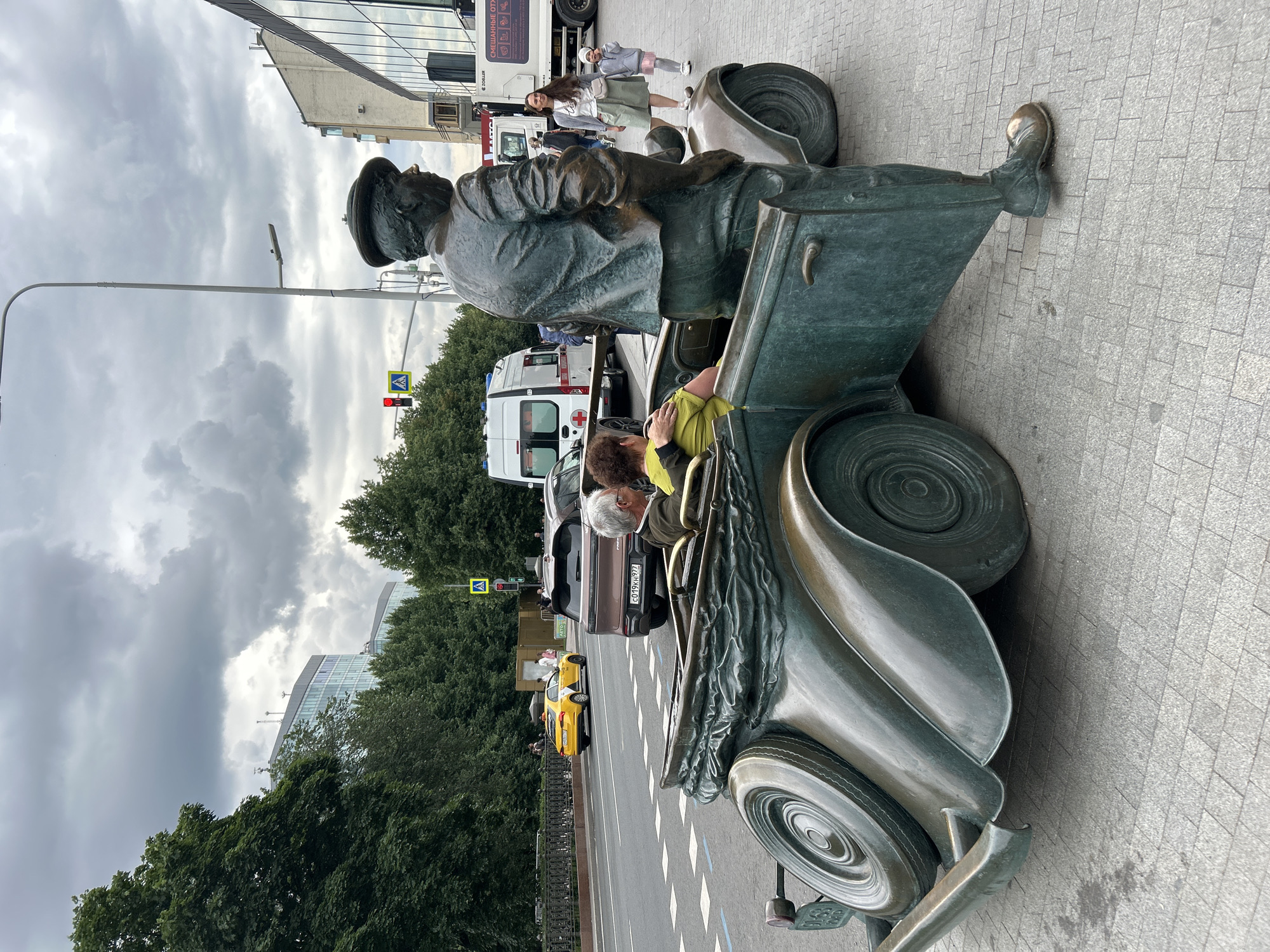 The image depicts a bronze sculpture of a man in a cap leaning against an open car door of a vintage convertible. The sculpture is situated on a pedestrian walkway paved with gray stone tiles. In the background, a street with traffic is visible. Cars, including a yellow taxi, a brown SUV, and an ambulance, are present. Several people, including a woman and a child, are walking on the walkway. A multi-story building with a modern glass facade stands behind them. The sky is overcast with heavy clouds. This sculpture is located in Moscow, Russia, specifically at the intersection of Tverskaya Street and Okhotny Ryad Street, near the State Duma building.