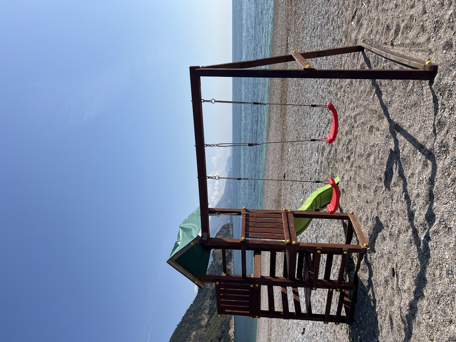 A wooden children's playground structure is positioned on a pebble beach. The structure, predominantly dark brown, features a playset on the left comprising a ladder, an elevated platform with a green tarp roof, and a green plastic slide extending to the ground. Yellow plastic caps are visible on various structural points. To the right, an attached swing set holds two swings with red plastic seats suspended by dark ropes. Distinct shadows from the playground are cast onto the light-colored pebbles of the beach, indicating bright sunlight from an overhead or slightly angled source. Behind the playground, tranquil blue-green ocean water extends towards a distant horizon. A large, verdant mountain or hill occupies the left background, sloping down to meet the sea. Further in the background, across the water, additional landmasses or mountains are faintly visible under a clear, light blue sky. No individuals are present in the scene. The location is Unknown, Unknown.