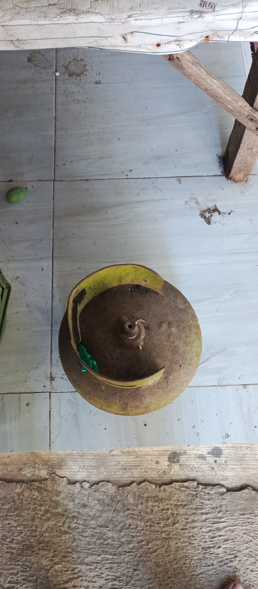An overhead shot captures a rustic outdoor scene in Arjasa, Indonesia, likely a backyard or covered patio. The ground is tiled with light gray square tiles, some showing signs of wear and dirt. In the center, a weathered, olive-green LPG gas cylinder rests on its side. Its top valve mechanism is clearly visible, and a piece of bright green plastic is tucked into its rim. To the left of the cylinder, a small, light green object, possibly a piece of fruit or vegetable, lies on the tiles. In the background, rough-hewn wooden planks form a simple, unfinished structure. A small brown smudge on the tiles to the left of the cylinder suggests a recent spill or stain. The overall impression is one of everyday utility in a simple, rural setting.