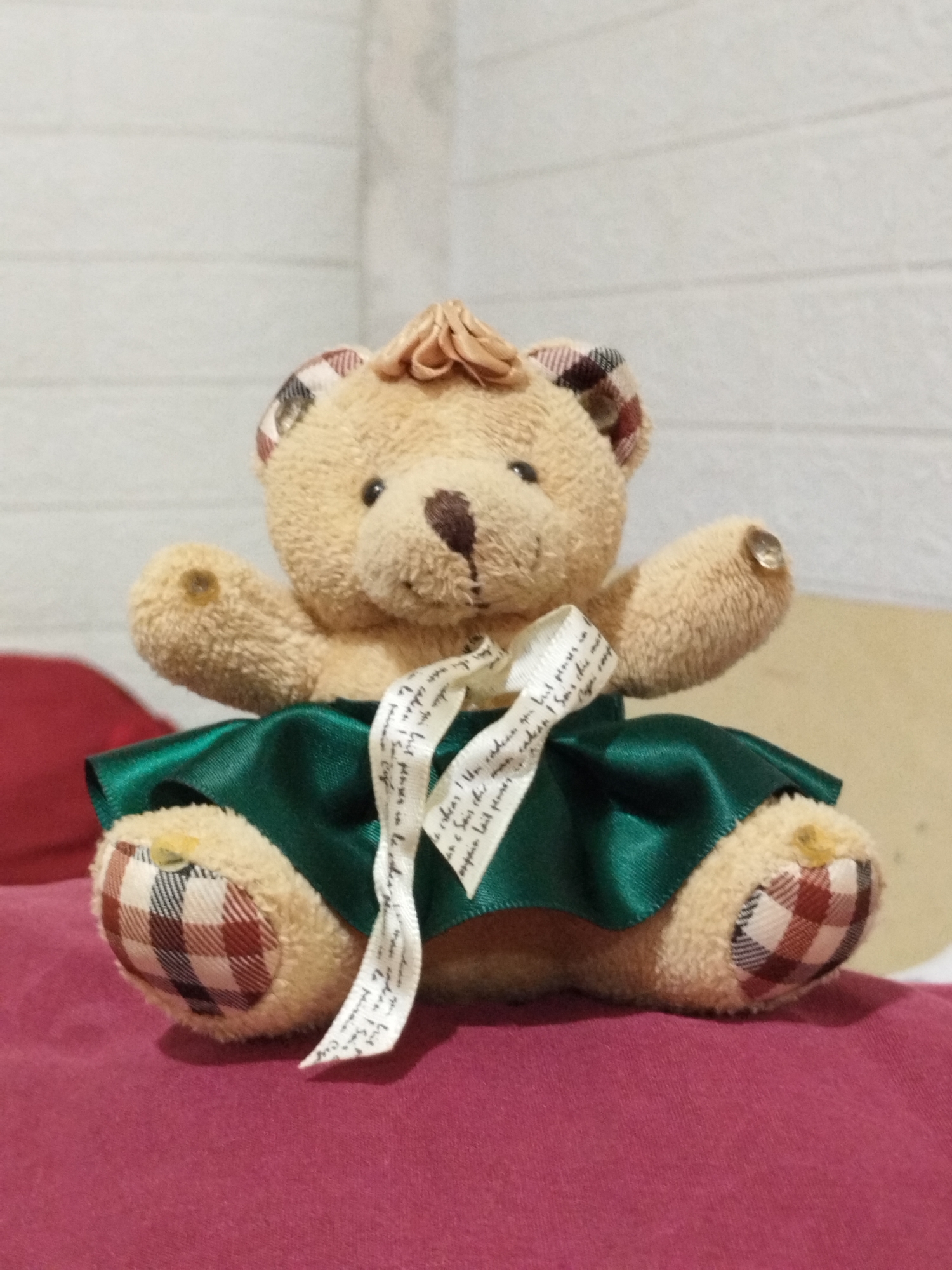 A teddy bear is seated on a red surface. It has a tan body, plaid ears and feet, and a brown nose. The teddy bear is wearing a green skirt tied with a white ribbon. The background consists of a white, textured wall. The location is unspecified, but it's requested it should be mentioned that the place is in Lhokseumawe, Indonesia.