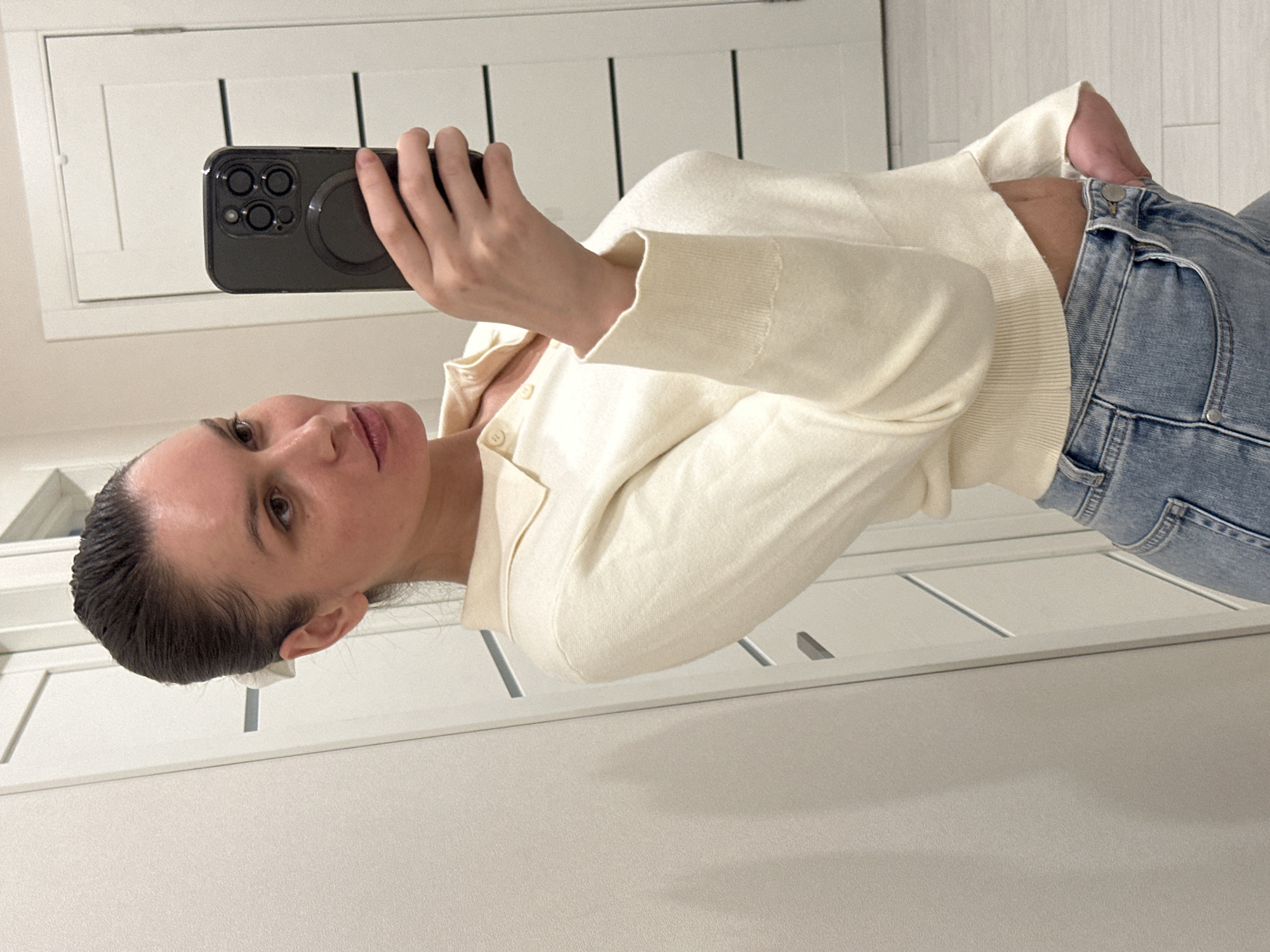 A woman is captured in a mirror selfie in an indoor setting, possibly in Khimki, Russia. The image is oriented sideways, rotated 90 degrees clockwise. She holds a black smartphone in her right hand, featuring a prominent triple-lens camera system and a circular magnetic accessory on its back. She appears to be looking at the phone's screen or camera in the reflection.
The woman has dark hair pulled back from her face. She is wearing a cream or off-white long-sleeved top with a collared neckline and a few buttons down the front, paired with light blue denim jeans. A small portion of her midriff is visible between her top and jeans. Her expression is neutral.
The background, visible as a reflection, consists of white, paneled doors or cabinets, suggesting a modern interior space like a bedroom or hallway with built-in storage. The lighting is soft and even, consistent with indoor conditions. There is no visible text in the image.