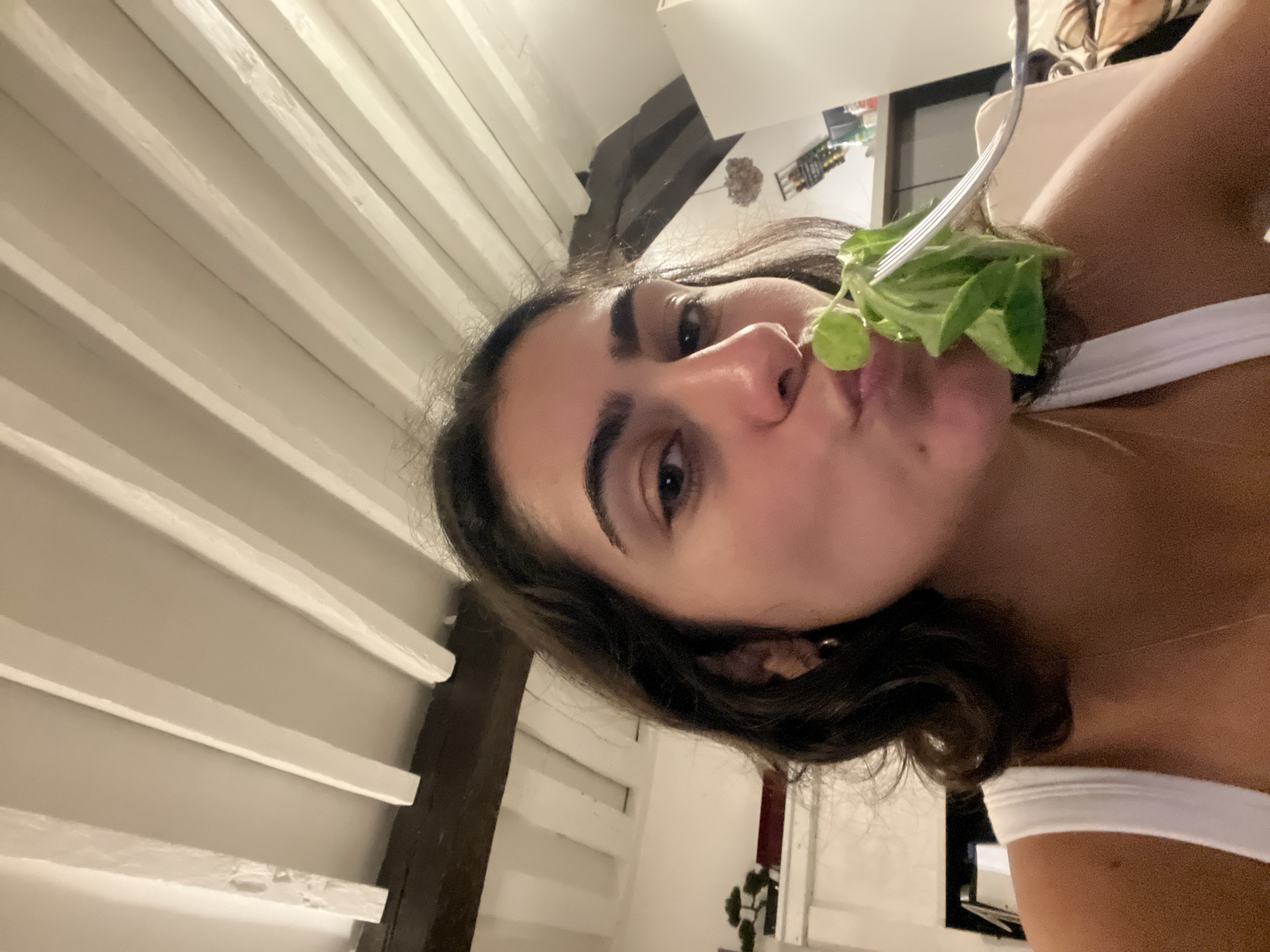 A person with long dark hair is holding a fork with lettuce near their mouth. They are wearing a white top. The background includes white ceiling beams and a glimpse of a shelf unit with various objects. It is impossible to determine the location in Paris, France, from this image.