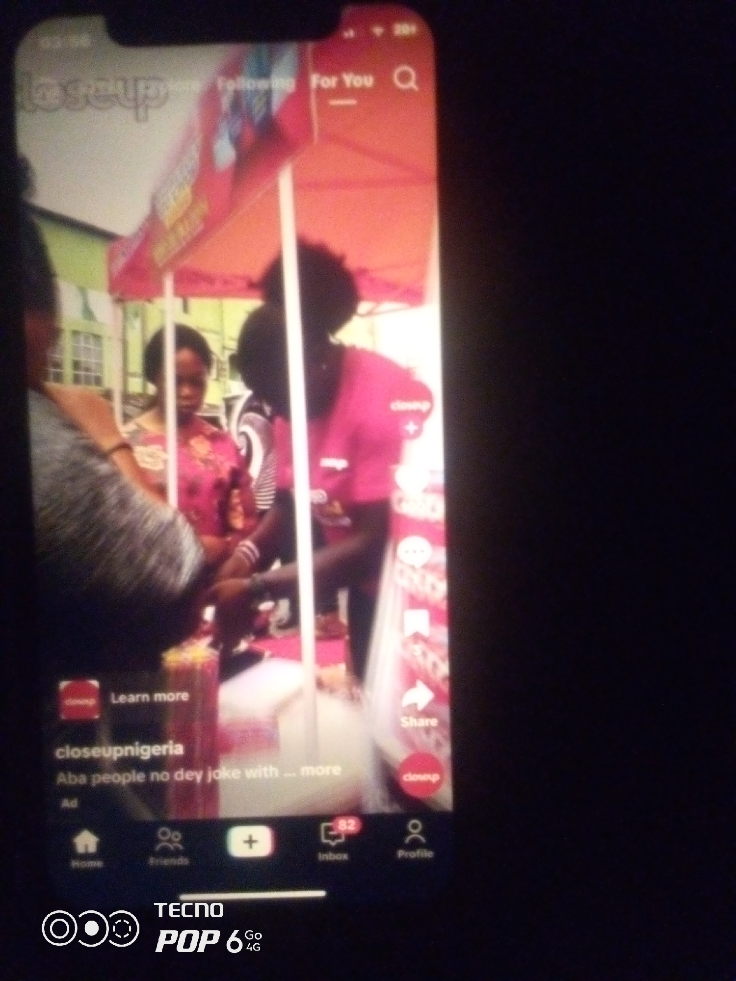 The image shows a smartphone screen displaying a social media video. The video appears to be a short advertisement or a user-generated clip featuring people under a red and orange canopy. The canopy has branding that is partially visible, including the word "closeup" and possibly a logo. In the foreground of the video, a person in a pink shirt is visible, and to their left, another person in a patterned pink and white shirt is also seen. The lower part of the phone screen shows social media interface elements, including icons for home, friends, a plus sign for creating content, and an inbox with a notification count of 82. The username "closeupnigeria" is displayed, along with the text "Aba people no dey joke with ... more". The Tecno Pop 6 4G logo is also visible at the bottom of the phone screen. The image is blurry and taken in low light conditions, making it difficult to discern specific details about the people or the exact location. However, the text "closeupnigeria" and "Aba people" suggest a connection to Nigeria, and potentially the city of Aba within Nigeria. The context of the video within the social media app implies it is a digital representation of an event or product demonstration rather than a direct depiction of a specific physical location in Howayi, Nigeria.