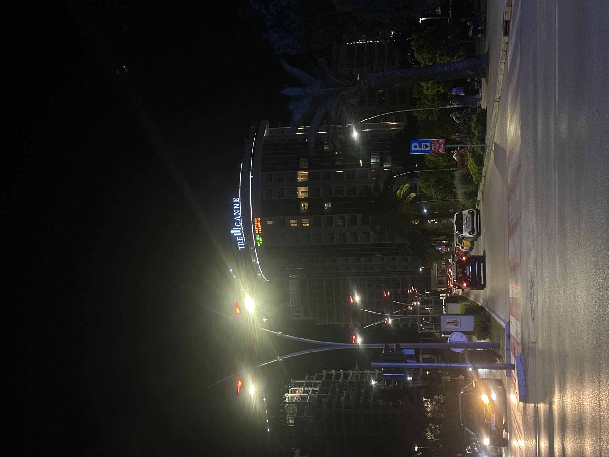 The image shows a nighttime scene in Budva, Montenegro, featuring the Tre Canne hotel. The tall, modern building is illuminated with its name displayed vertically. A palm tree stands beside the hotel. Streetlights line the avenue, and traffic is moving along the road, including cars with headlights visible. Traffic signals are visible. A parking sign is present on the sidewalk. The sidewalk is adjacent to the street and separated by a curb painted with red and white stripes. Further down the street, there appears to be a construction site. The sky is dark.