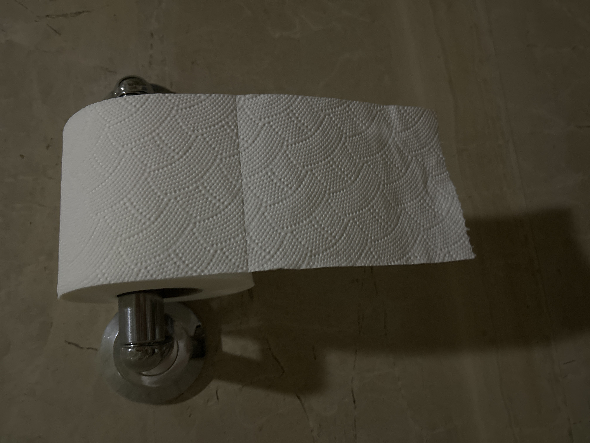 A roll of white toilet paper is mounted on a chrome holder on a light brown wall with a marble-like texture. The toilet paper is partially unrolled, with some sheets hanging down. The scene is dimly lit, casting shadows on the wall behind the toilet paper. The location is not evident from the image.