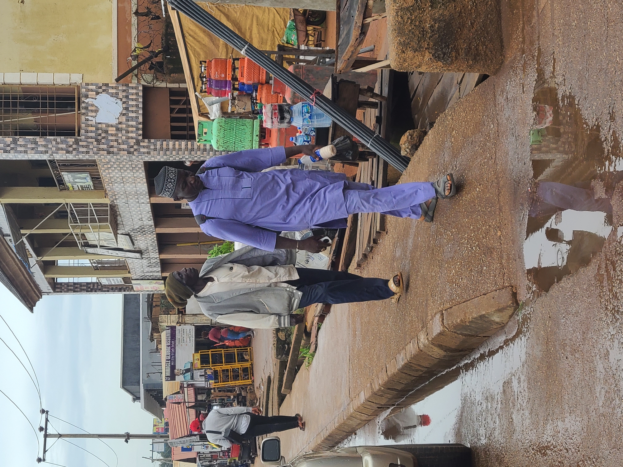 The image depicts two men walking on a sidewalk beside a street in what appears to be a busy urban area of Unknown, Unknown. The sidewalk is wet, reflecting the surrounding environment. The man on the left wears a gray jacket, dark pants, and sandals, while the man on the right wears a lavender robe and a black and white skullcap. The man on the right carries a bundle of long black pipes. Behind them is a commercial district with buildings, signs, and parked vehicles. A sign for "MIRACLE GLOBAL ASSEMBLY" is visible on one of the buildings. Various items like propane tanks and stacked containers are visible behind the men, suggesting a commercial or industrial presence. Overhead power lines run along the street.