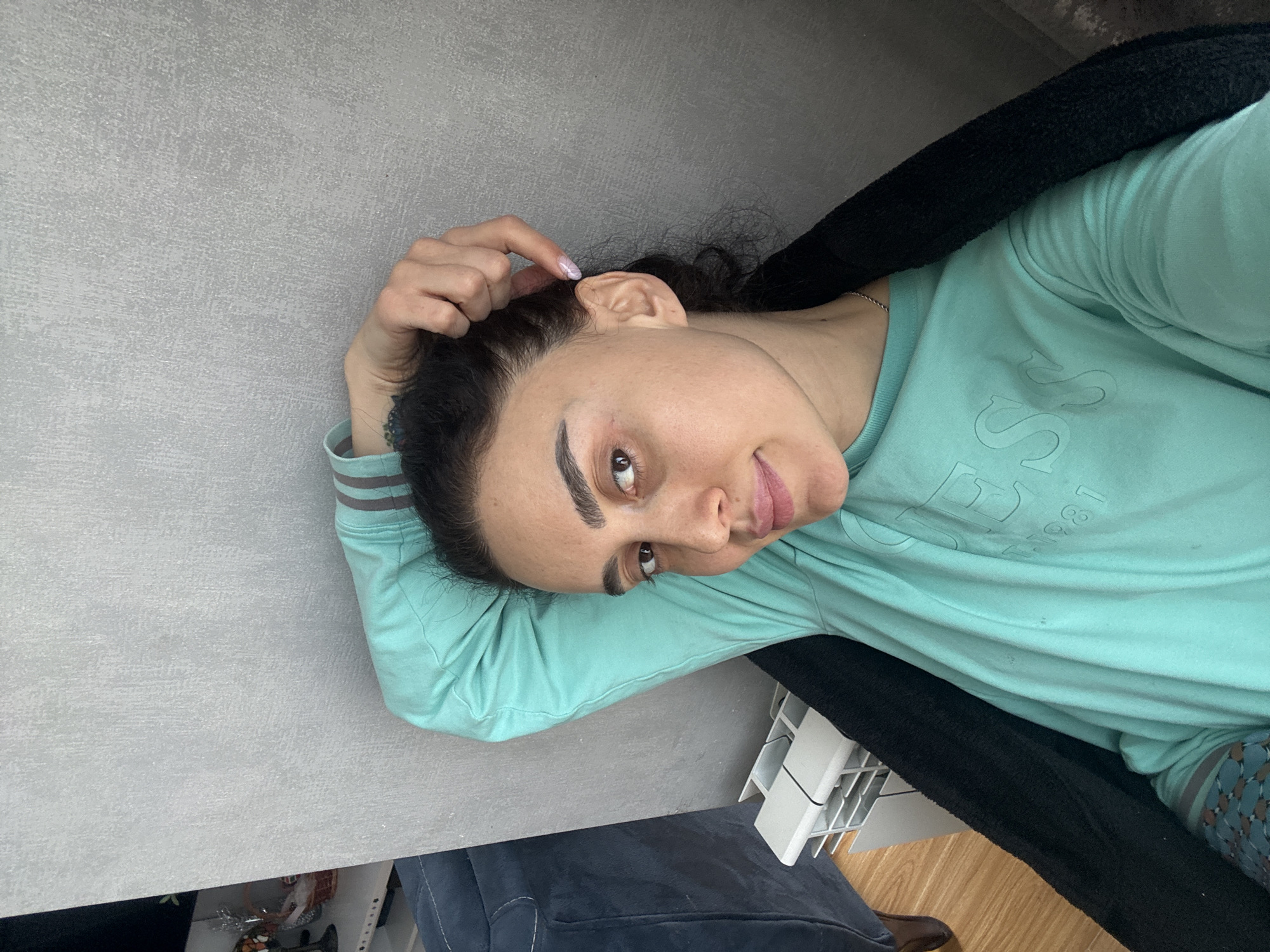 A woman with dark hair is lying at an angle against what appears to be a gray countertop. She is wearing a light turquoise long-sleeved shirt with gray stripes on the sleeves and a black garment draped around her neck. Her head rests near her left hand. The background includes parts of a blue upholstered item, a white plastic structure, and a floor with a wooden finish. The location within Tbilisi, Georgia, cannot be determined from the image.