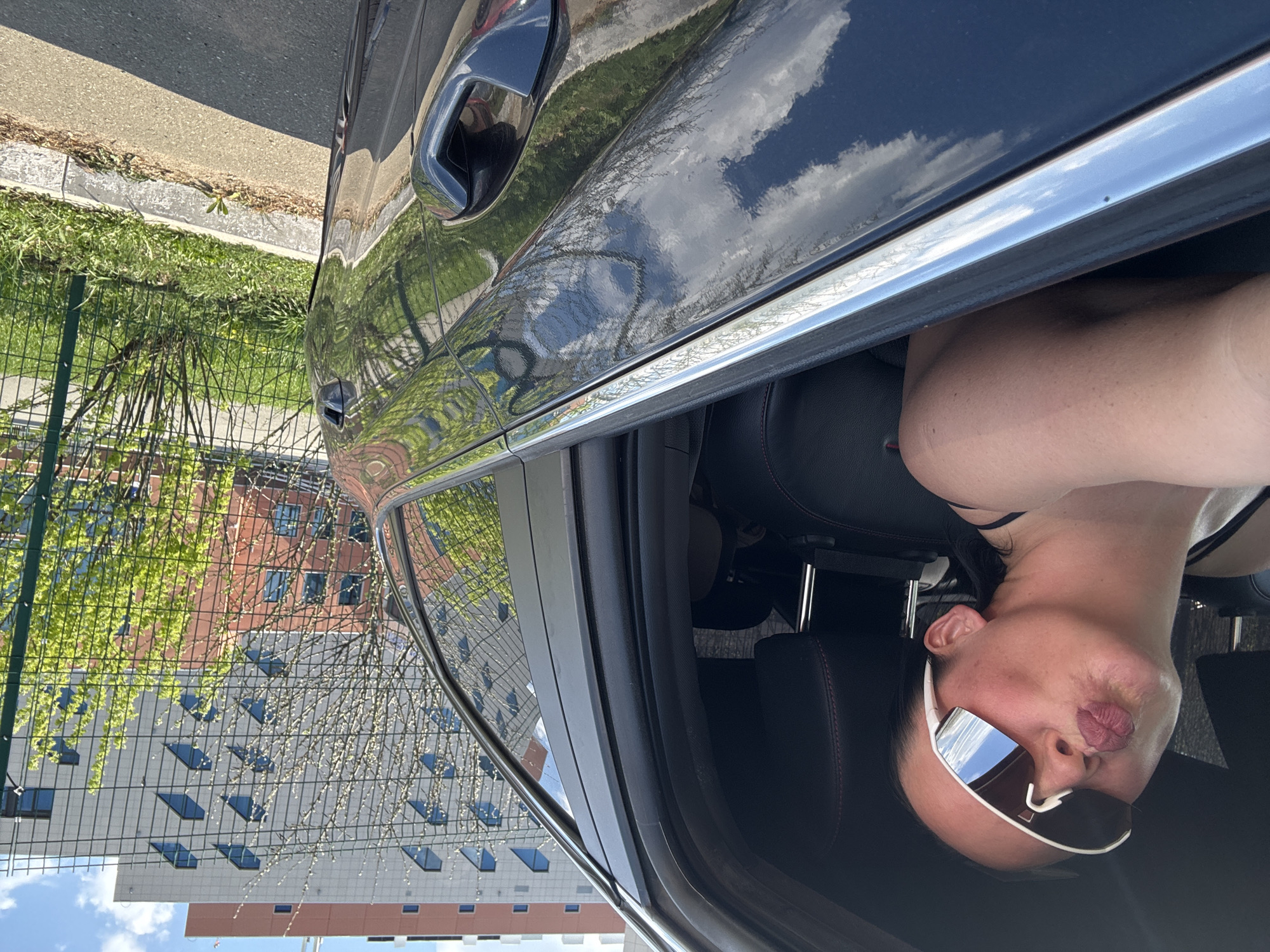 The image shows a woman wearing sunglasses sitting inside a car. The car door is open, and the reflection in the car's paint shows a building and a wire fence with greenery behind it. Based on visual cues, this image was likely taken in Прикамский, Russia.