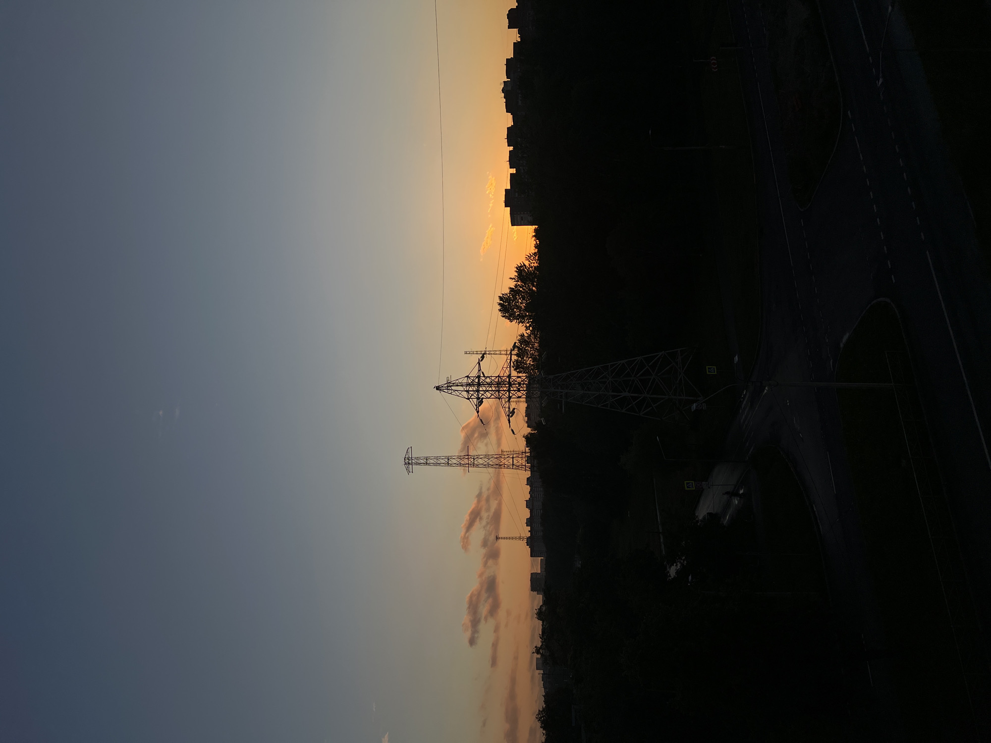 This is a vertical, medium-distance, silhouette photograph of an urban landscape at dusk. The sky is a gradient of deep blue at the top, transitioning to a pale yellow and orange near the horizon, indicating the setting sun. A few wispy, illuminated clouds are present in the orange band. Silhouetted against the sky are several elements of the city. In the mid-ground, the dark outlines of apartment buildings form a skyline. In front of the buildings, a dense band of dark trees is visible. Dominating the foreground and mid-ground are the silhouettes of two large electricity pylons, with the one on the right being closer and larger. A network of power lines connects between them and extends further into the scene. Below the pylons, a dark, tree-lined area gives way to a road intersection with streetlights. The road surface appears wet, suggesting recent rain. The scene is devoid of people. The image captures a quiet, industrial edge of Saint Petersburg, Russia, during the twilight hours.
