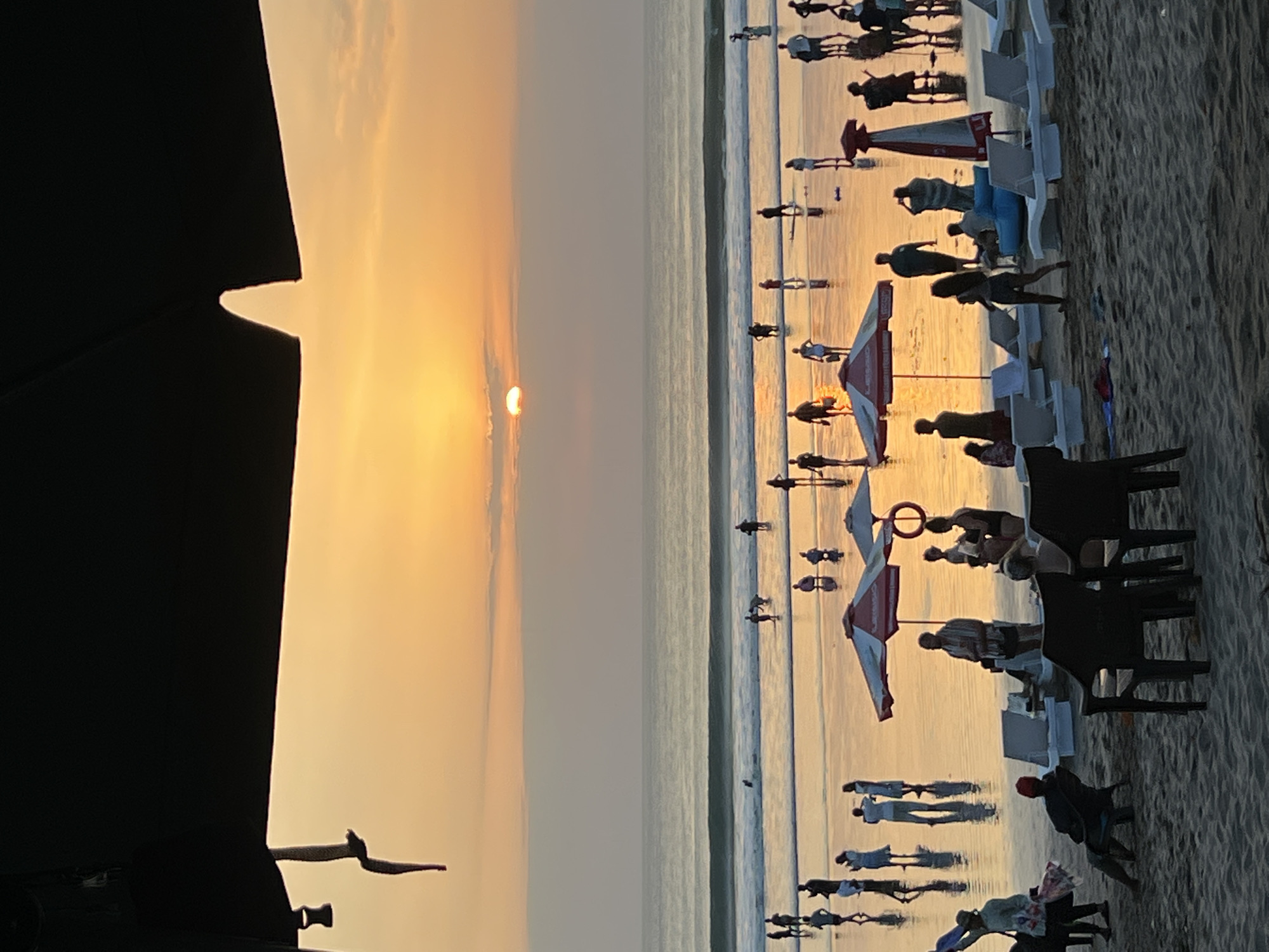 The image depicts a beach scene in Kuta, Indonesia during sunset. The sky is painted with hues of orange and yellow, with the sun partially obscured by clouds. The ocean is calm with gentle waves, reflecting the warm colors of the sky.
On the beach, numerous people are visible. Some are walking along the shore, while others are standing in the shallow water. Several individuals are seated on beach chairs, and there are also beach umbrellas providing shade. In the foreground, there are beach chairs and umbrellas arranged on the sand.
The scene suggests a relaxed atmosphere, with people enjoying the end of the day by the sea. The overall impression is one of a popular tourist destination, likely Kuta Beach, known for its scenic sunsets and vibrant atmosphere.