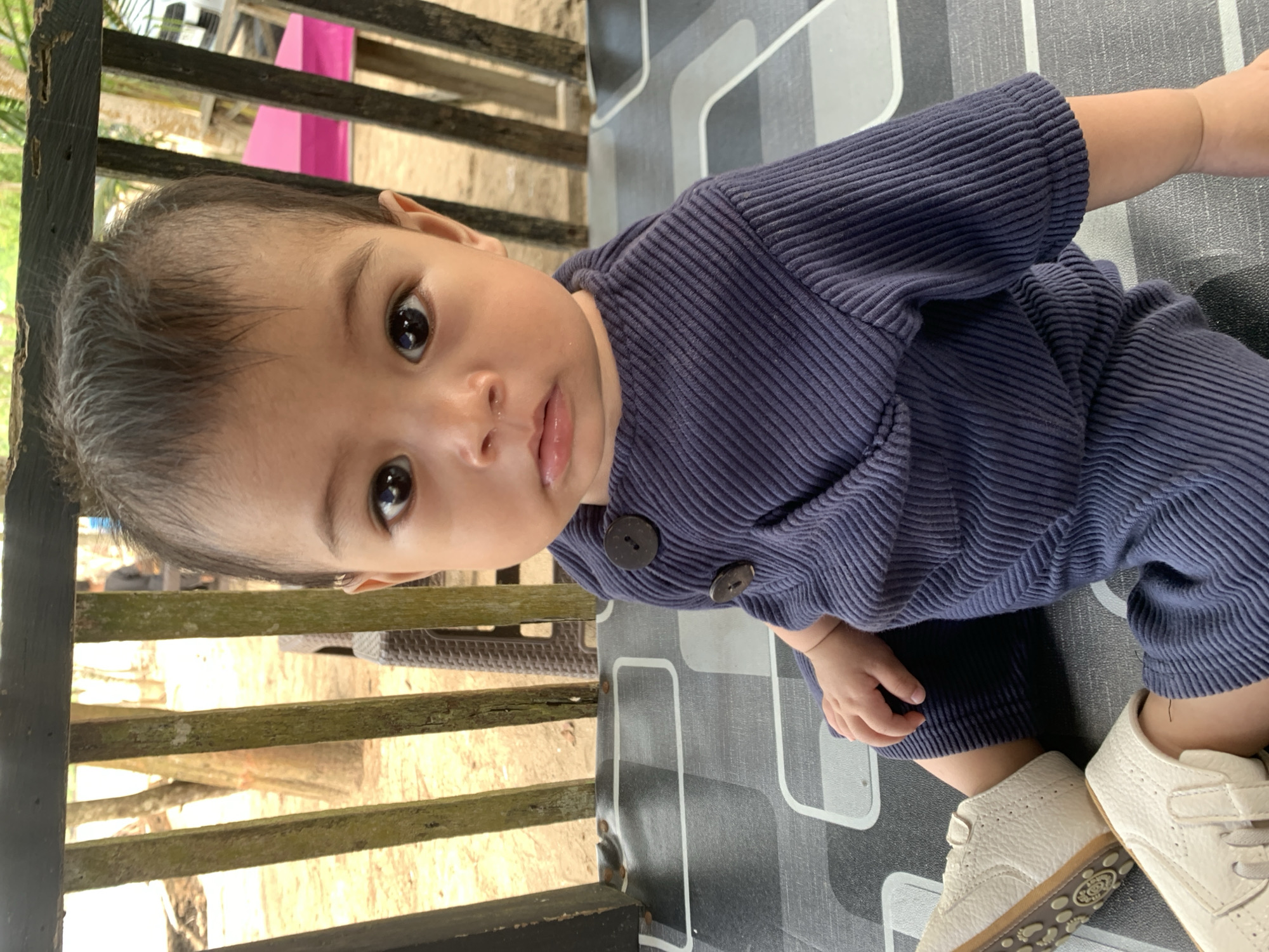 In Tanjung Limau, Indonesia, an infant is pictured wearing a navy-blue, corduroy-ribbed outfit with dark buttons. The child is wearing cream-colored leather shoes. The baby is sitting on a grey patterned surface. In the background is a wooden structure. The photo is taken from an unusual perspective.