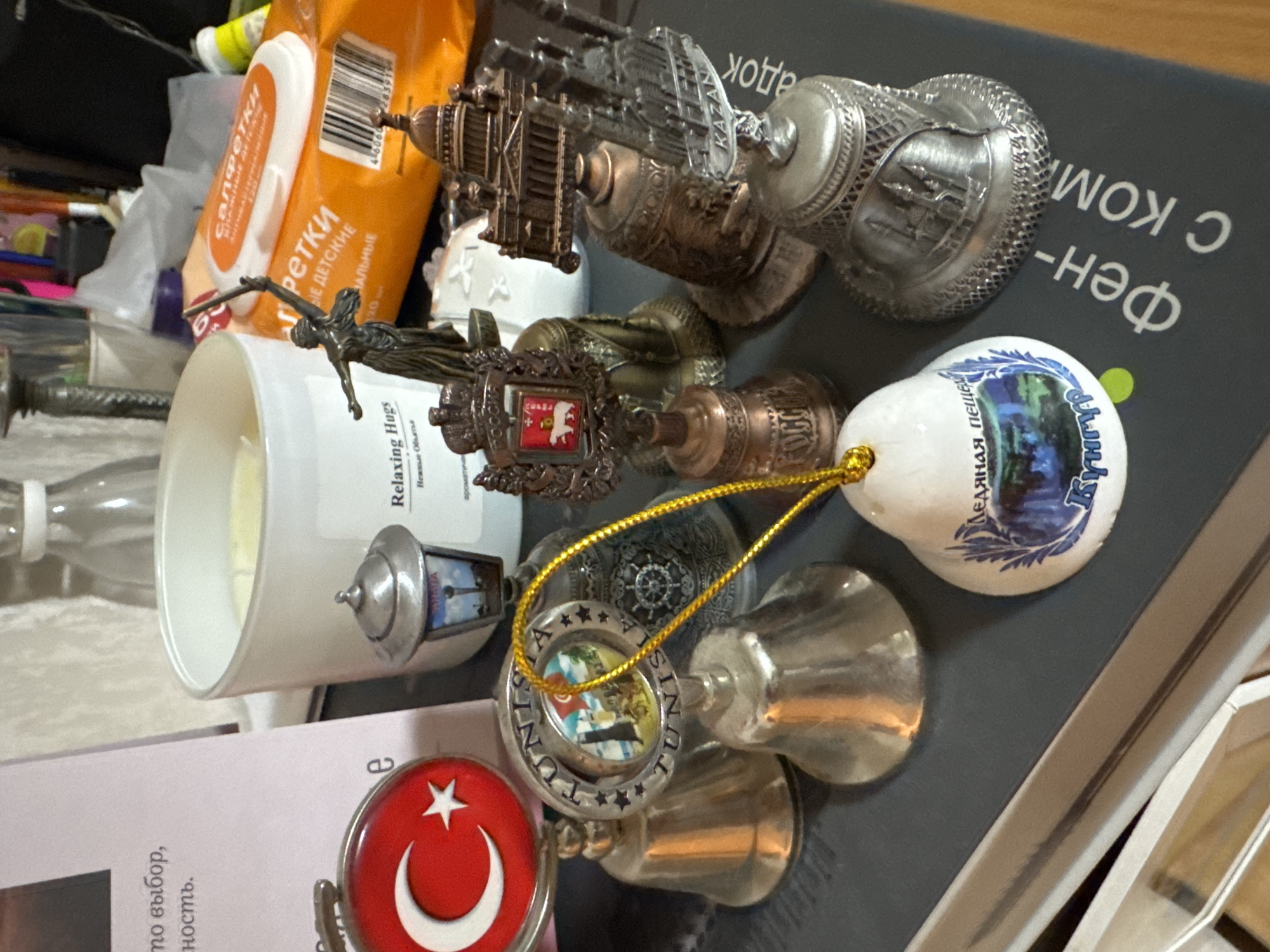 The image depicts a collection of souvenir objects on a surface with Russian text. These souvenirs include miniatures of notable structures and landmarks from various locations. Specifically, a representation of the Kul Sharif Mosque in Kazan, Russia, is visible, along with a souvenir bell bearing the image of the Kungur Ice Cave. The Turkish flag is also seen. Other objects in the scene are a box of wipes, a candle, and miscellaneous items. The scene appears to be indoors.
