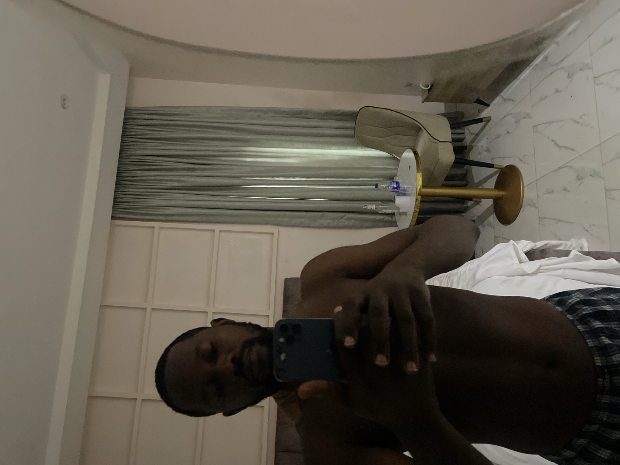 A shirtless man with dark skin, a beard, and short hair is taking a mirror selfie in a hotel room in Abuja, Nigeria. He is holding a smartphone in his hands, and his torso and arms are visible. He is wearing checkered boxer shorts. The room has light pink walls with a patterned design, grey curtains, a gold-legged table with a chair, and a marble-tiled floor. A bottle of water and tissue rolls are on the table. The mirror reflects the man, the room, and a glimpse of the bed.