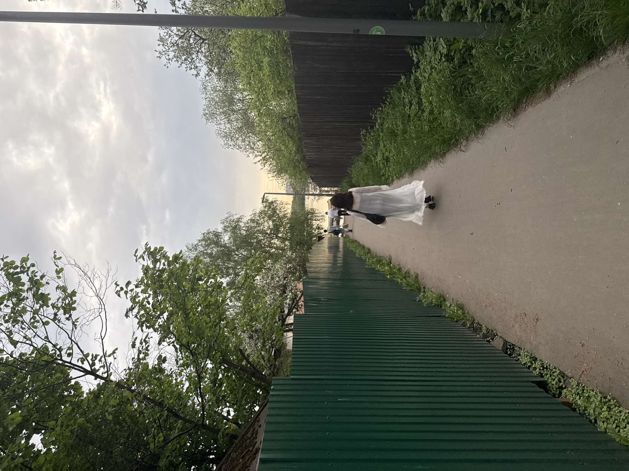 The image shows a scene in Moscow, Russia. A woman wearing a long white dress is walking along a paved pathway, with a black handbag on her shoulder. In the background, other people are also walking. A green, corrugated metal fence runs alongside the pathway on one side, while a brown wooden fence is visible on the other. Trees and vegetation are present on both sides of the path. The sky is overcast. The photo is taken from an angle, with the bottom of the frame capturing the green metal fence close up, and the pathway leading into the distance.