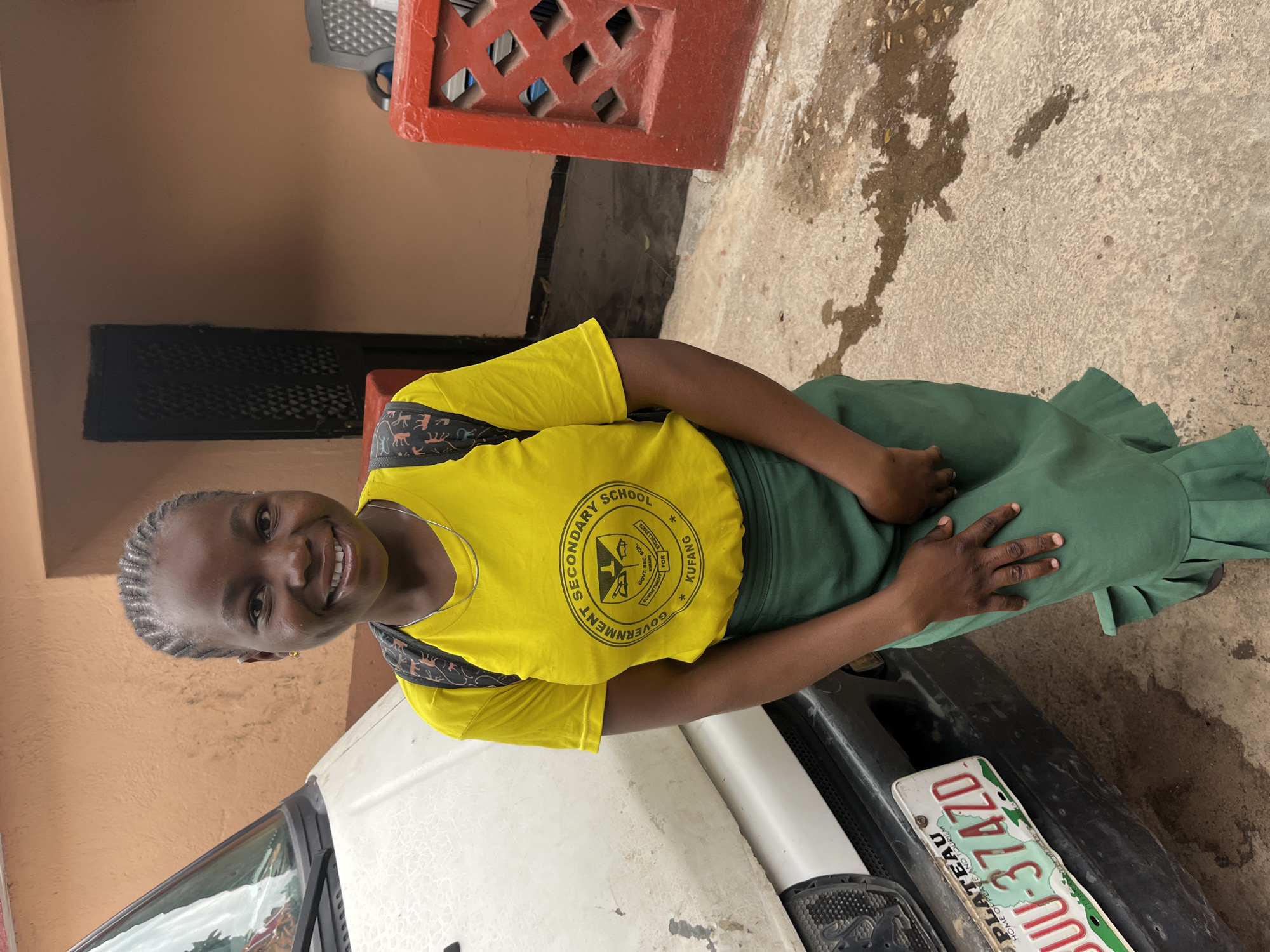 A young girl, appearing to be a student, is captured in a bright, outdoor portrait. She stands facing the camera with a wide, joyful smile, showcasing her neat braids and an earring. She is dressed in a school uniform consisting of a vibrant yellow short-sleeved top and a long, dark green pleated skirt. A backpack with a patterned strap, featuring various animal silhouettes, is visible over her right shoulder.
The yellow top features a circular school emblem with text clearly visible: "SECONDARY SCHOOL", "GOVERNMENT", "GOVT. SEC. SCH.", "KUFANG", and "COMMITMENT FOR EXCELLENCE".
The girl is positioned next to a white vehicle, possibly a car or van, which occupies the lower foreground of the image. Part of its license plate is visible, reading "PLATEAU" and "HOME OF PEACE AND TOURISM", along with the alphanumeric code "UU-374ZD". These details strongly suggest the location is Plateau State, Nigeria, consistent with Jos.
The background features a reddish-brown painted wall and a section of a red, decorative concrete block with square cutouts, possibly part of a fence or building facade. The ground is a light-colored, possibly concrete or paved surface, with darker, wet-looking patches. The lighting indicates it is daytime. The overall impression is one of a cheerful student in her school environment.