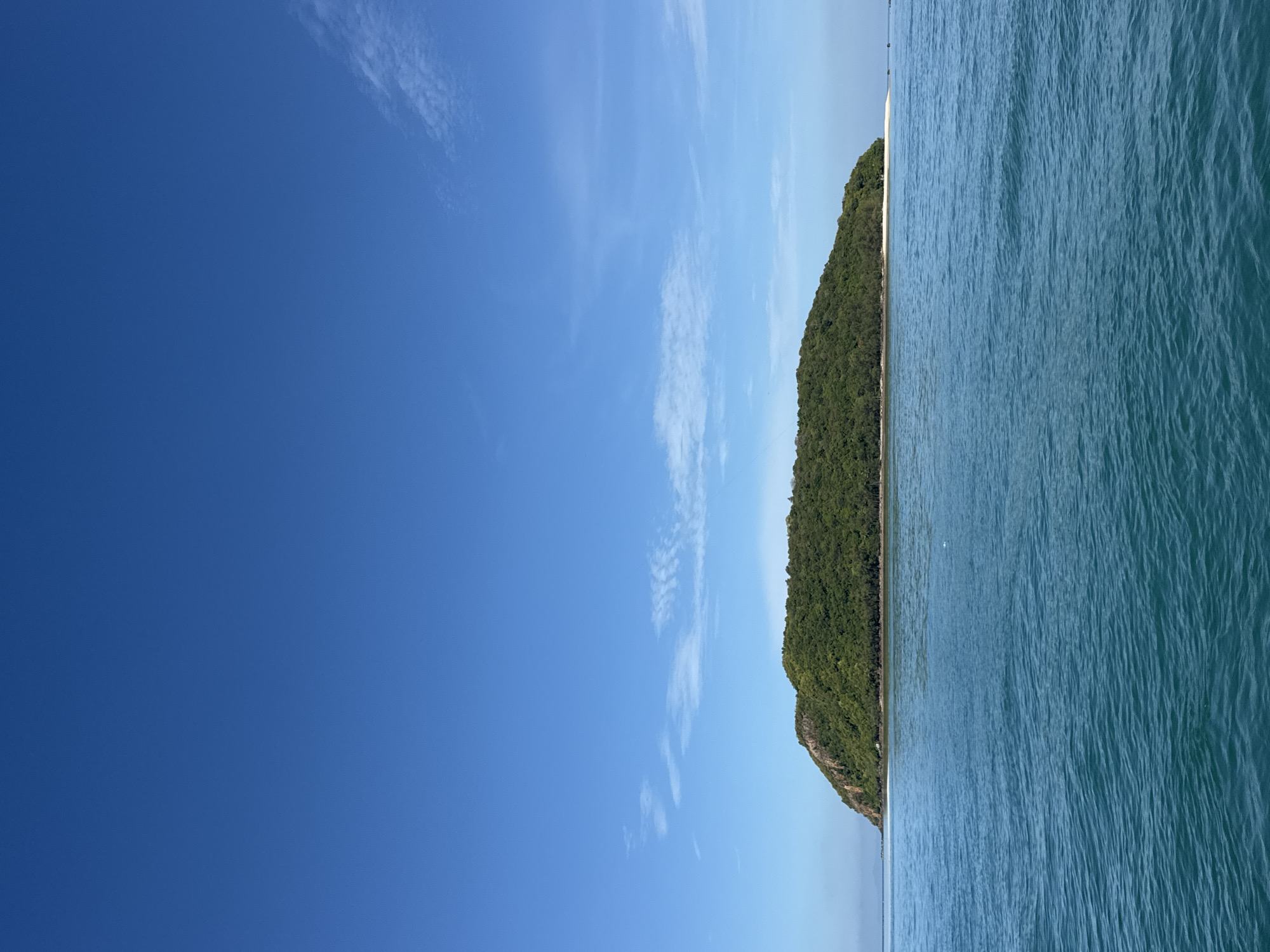 A wide shot shows a verdant, oval-shaped island in the middle distance, with a sandy beach visible on its right side. The island is covered in lush green trees and foliage, with rocky outcrops on its left side. The ocean in the foreground is a deep blue-green with gentle ripples. The sky above is a clear, bright blue with a few wispy white clouds. The scene appears to be a tropical paradise, likely in a coastal area. Given the location of ตำบลเกาะพะงัน, Thailand, this image could depict Koh Phangan, an island known for its natural beauty and beaches. There are no people or man-made structures visible in the image.