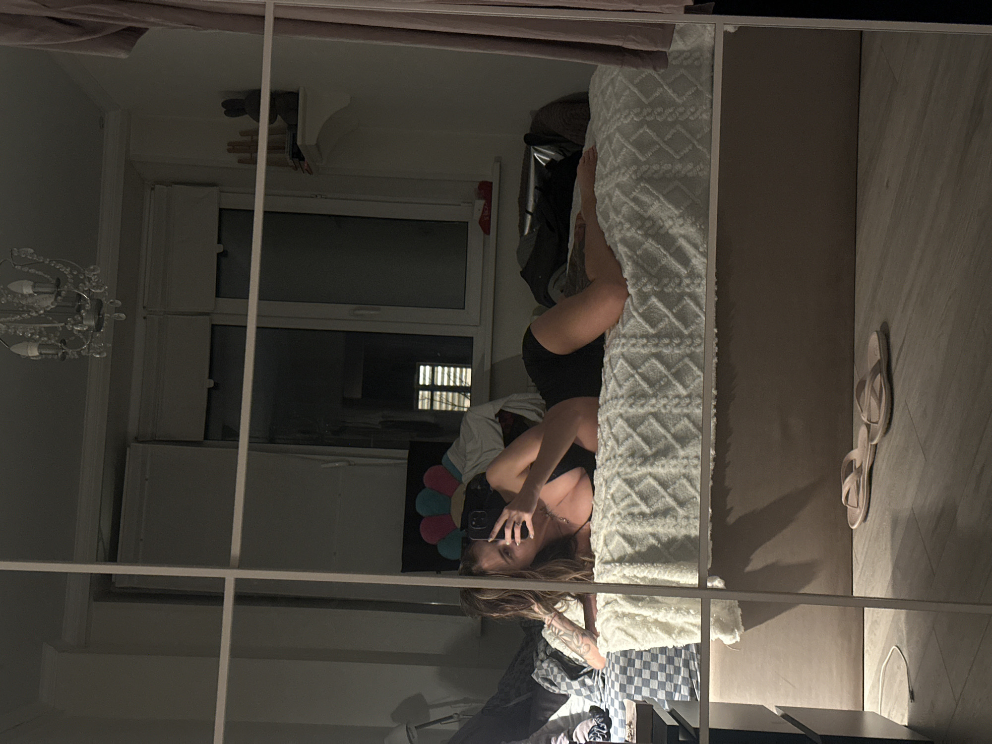 A young woman is lying on a bed, taking a selfie in a mirrored wardrobe. The room appears to be a bedroom in a residential apartment. The bed is covered with a white quilted blanket. The woman is wearing a black top and black shorts. She has long brown hair and tattoos on her arm. In the background, a window with blinds is visible, as well as a chandelier and a shelf with decorative objects. A pair of flip-flops are on the floor in front of the wardrobe. The reflection in the mirror also shows a corner of a dresser and a lamp. The overall lighting suggests it is nighttime or the room is dimly lit. No elements in the image suggest a specific location in Moscow, Russia.
