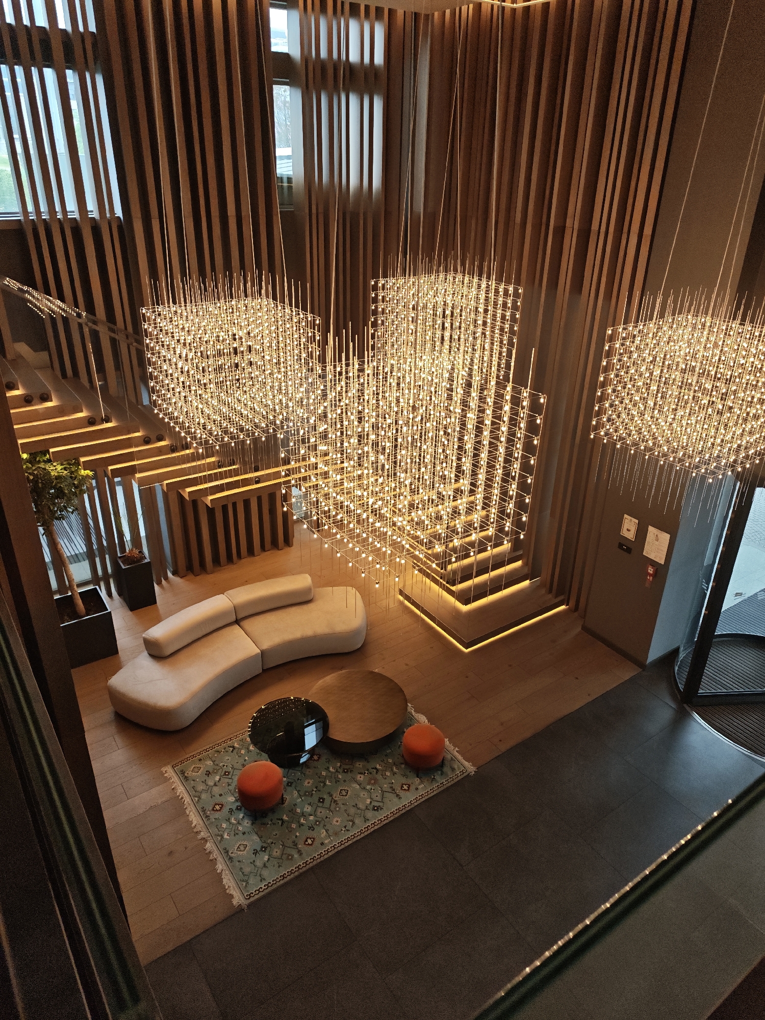 The image captures an interior view of a modern, stylish lobby, potentially in a hotel or office building, situated in Sofia, Bulgaria. The architecture is characterized by vertical wooden slats that adorn the walls and windows, providing a sense of height and texture.
The lobby features a staircase on the left side of the frame, illuminated by recessed lighting on each step. The staircase ascends, indicating the presence of multiple floors.
Below the staircase is a curved, light-colored sofa and a rug with two round coffee tables on it. Two orange poufs sit on the rug.
The space is lit by three large, geometric chandeliers composed of many small light points suspended from the ceiling. The light fixtures add warmth and a sense of luxury to the scene.
On the right side, a door leads to an exterior area. The floor transitions from wood to a dark gray or black surface, likely tile or stone.
The image does not depict any people.