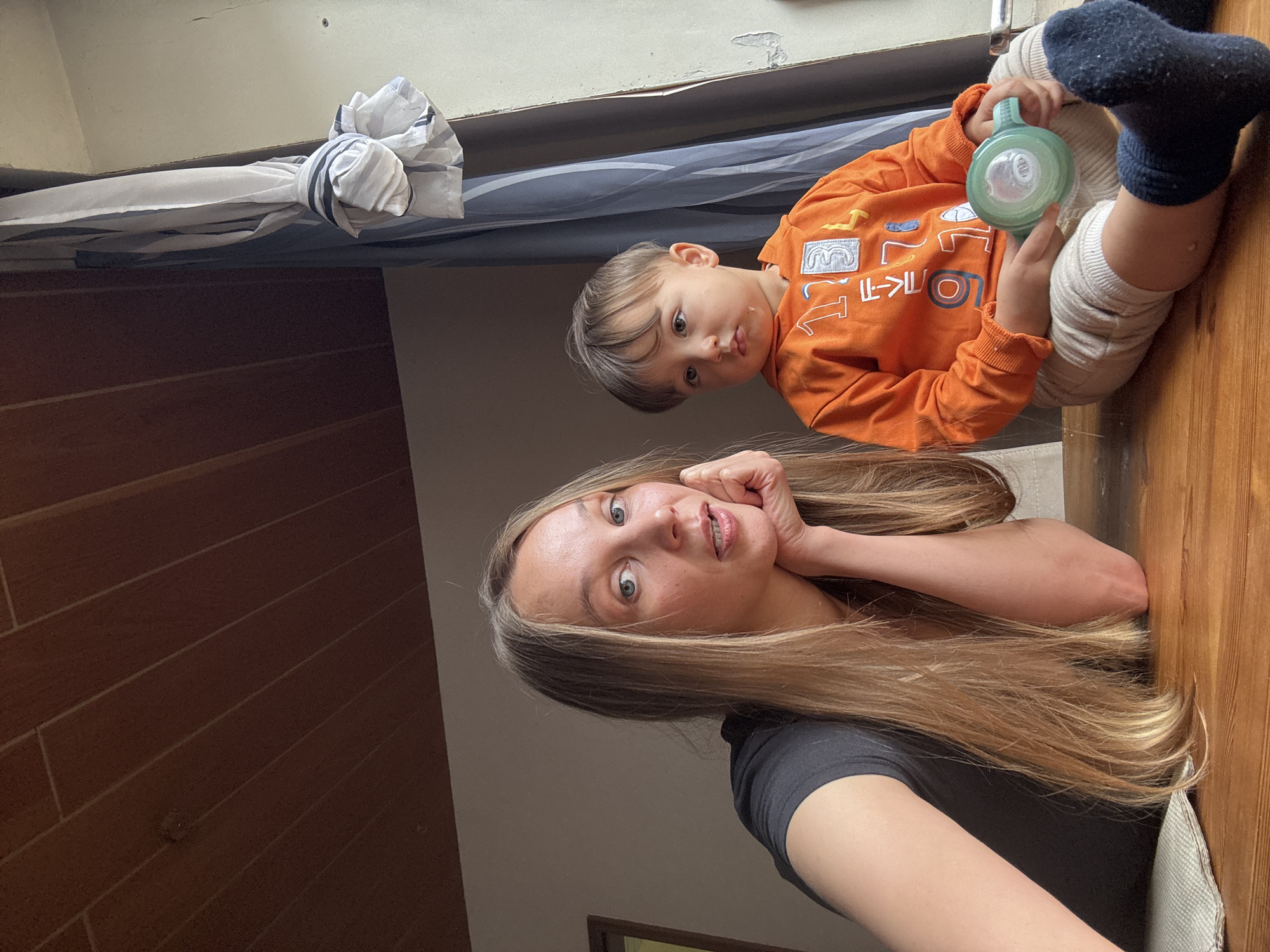 An eye-level, medium shot captures a blonde woman and a young child sitting at a wooden table. The woman, positioned on the left, leans on her hand, her gaze directed towards the camera. She has long, blonde hair and is wearing a black t-shirt. The child, on the right, wears an orange hooded sweatshirt and light-colored pants. The child is holding a green sippy cup and looking off to the side. The background features a wooden ceiling, a window with curtains, and a neutral-colored wall. This scene appears to be indoors, possibly in a residential setting in Überherrn, Germany.