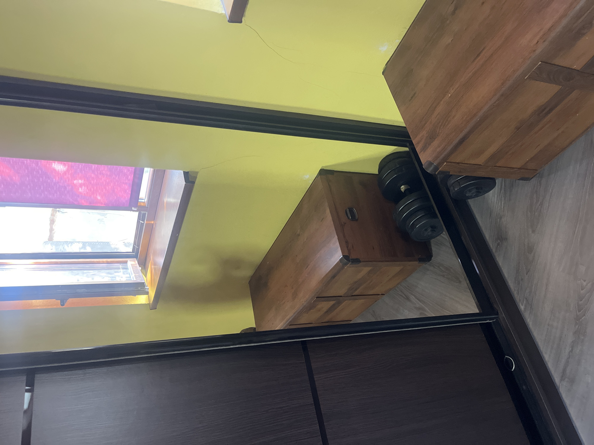 This is a photograph of an interior space, likely a room. On the left side of the image, a dark brown, wooden wardrobe with sliding doors is visible. A large mirror spans across the center of the image, reflecting the opposite wall. The wall is painted a bright yellow and features a window with a wooden sill. A purple roller blind is partially drawn over the window.

Reflected in the mirror are two wooden chests. The chest closer to the mirror has a dark, polished wood finish and appears to be a storage trunk with metal corner reinforcements. To the left of this chest, also reflected in the mirror, are two black dumbbells with circular weights. Another wooden chest, similar in style to the first, is partially visible in the lower right corner of the frame, in the real space, not a reflection. The floor is covered with light grey laminate flooring. There are a few faint cracks visible on the yellow wall.

The location is specified as Tarnivtsi, Ukraine.