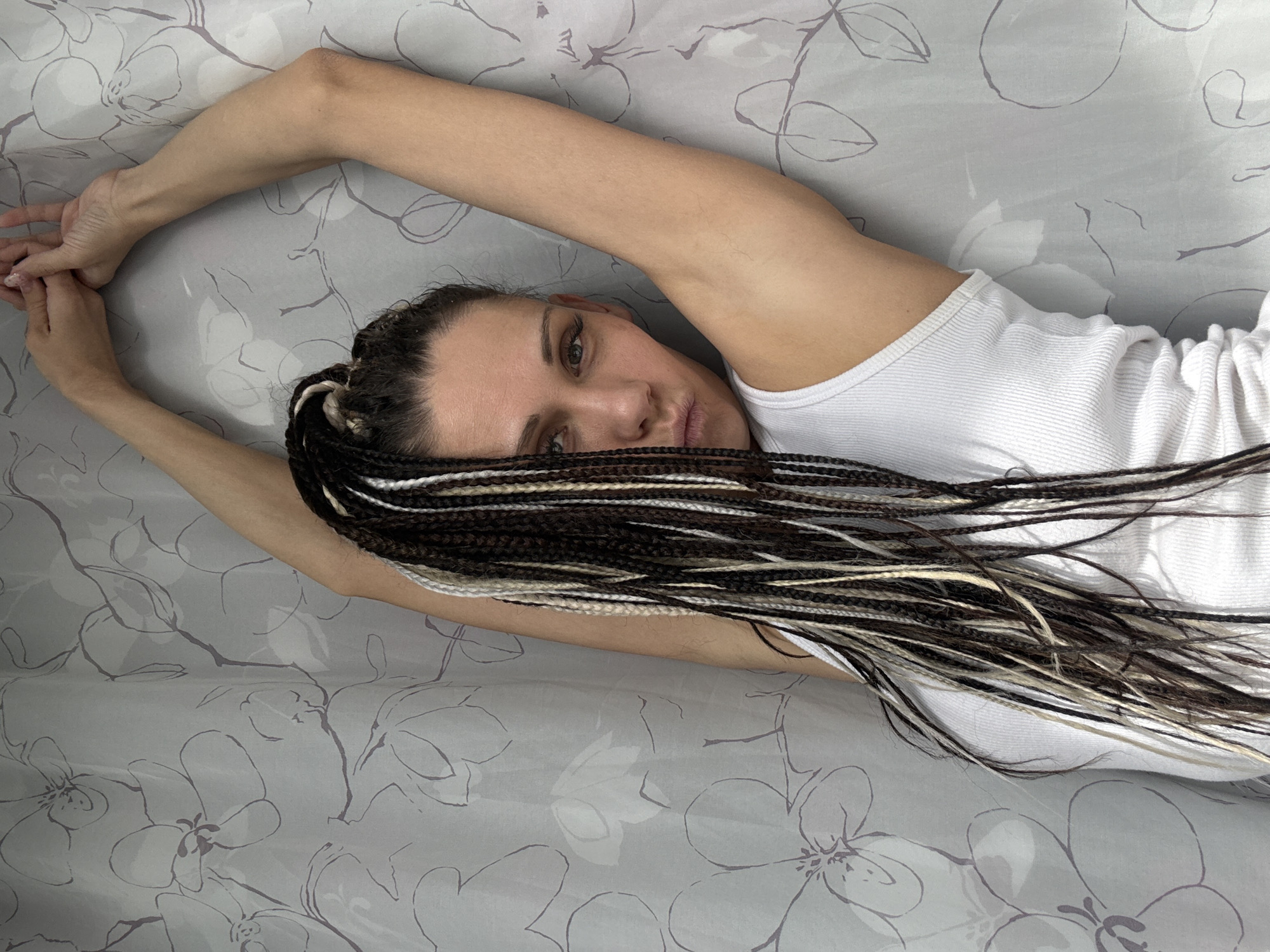 A woman is lying on a bed in Rusanovo, Russia. She is wearing a white tank top and has dark hair styled in braids with blonde highlights. Her arms are stretched above her head. The bed is covered with a gray floral patterned sheet.