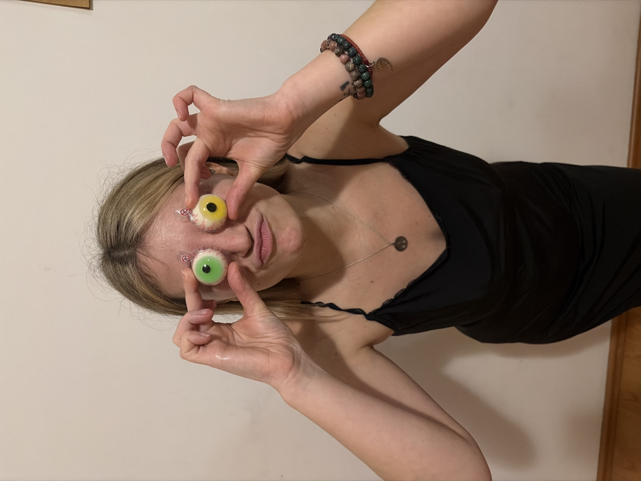 A woman holds two wrapped eyeball candies in front of her eyes. The candy on top is yellow, while the one below is green. She wears a black top with thin straps and a silver necklace. A bracelet adorns her left wrist, and a tattoo is visible on her forearm. The background is a plain off-white wall, and a wooden door is partially visible on the right. The image is rotated to the side.