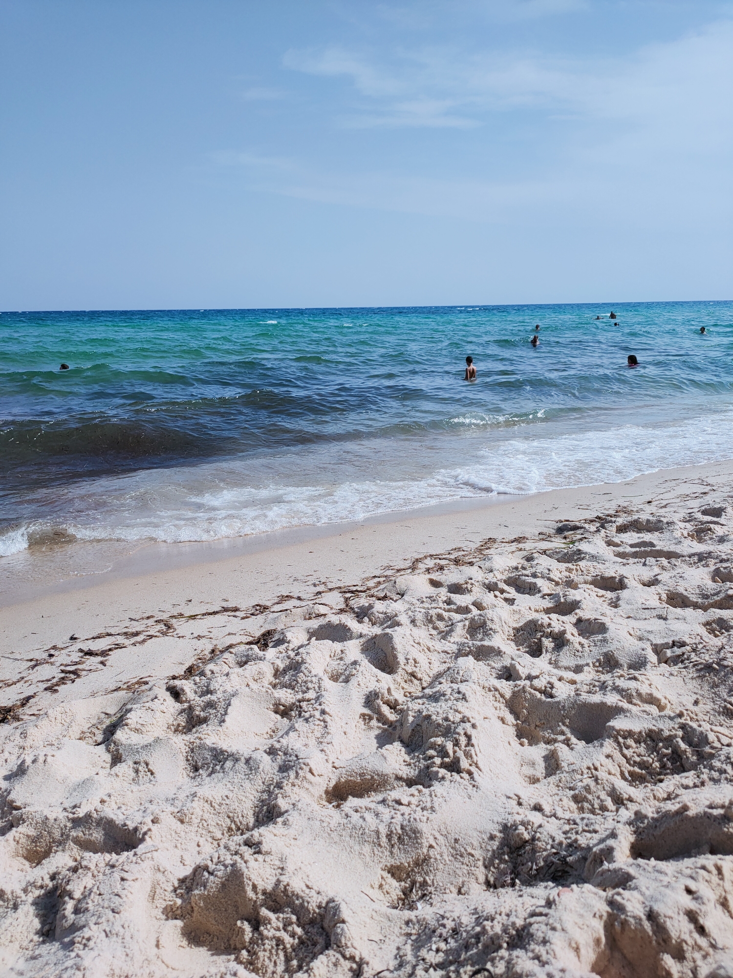The image captures a sunny day at a beach in Mrezga, Tunisia. The foreground is dominated by textured, light-colored sand with footprints and scattered brown seaweed. The midground shows the gentle waves of the Mediterranean Sea lapping at the shore, with white foam created by the breaking waves. The water transitions from a clear turquoise near the shore to a deeper blue further out. Several people are visible in the water, swimming or wading, with only their heads and upper torsos exposed. The background features a clear, pale blue sky with a few wispy white clouds. The overall scene is one of a peaceful beach environment with people enjoying the water on a bright, clear day.
