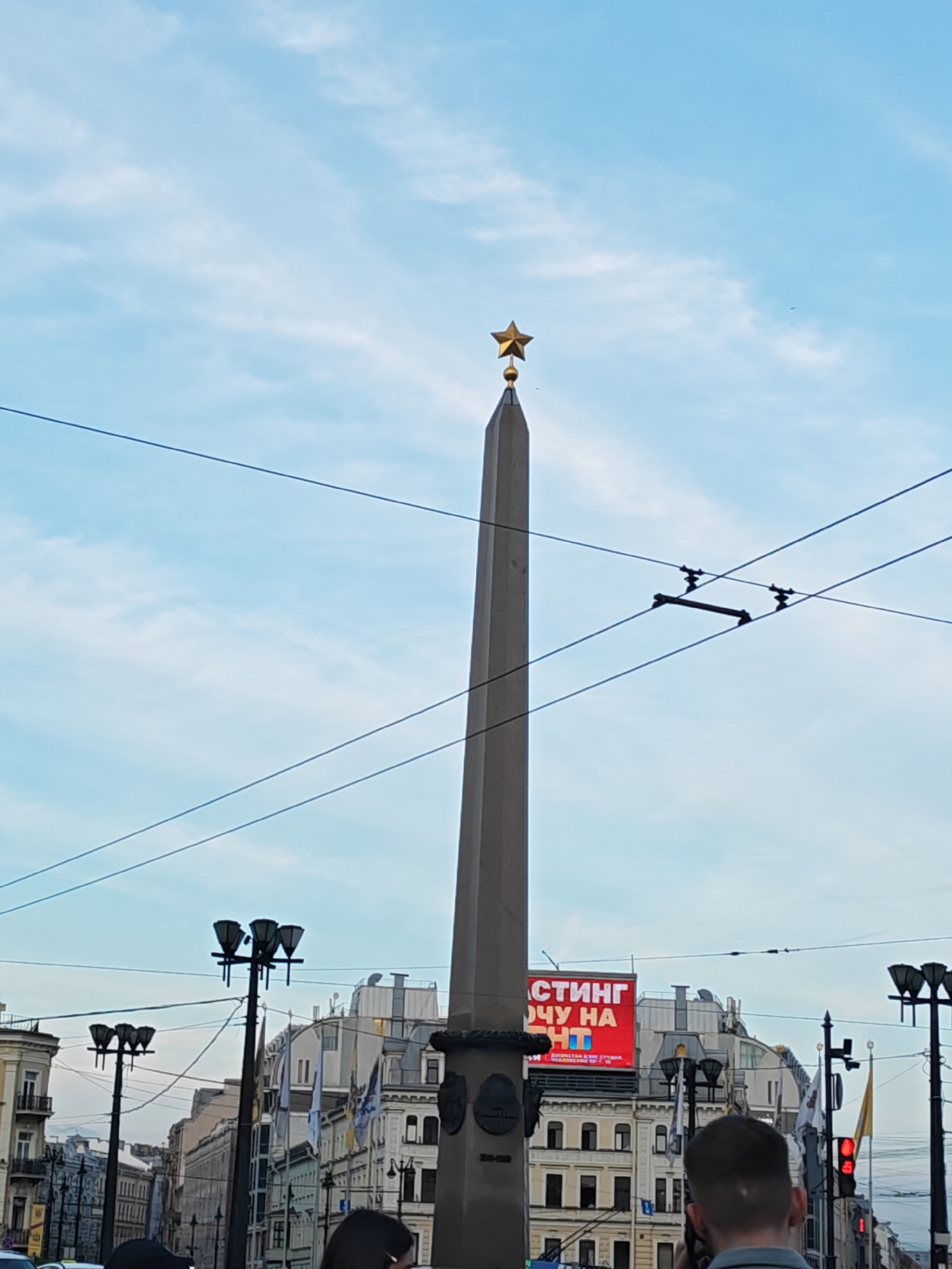 The image captures a ground-level view of the Obelisk of the Hero City of Leningrad (Saint Petersburg) in Vosstaniya Square, Saint Petersburg, Russia, under a light blue sky with scattered white clouds. The tall, grey, tapered obelisk stands centrally, topped with a prominent golden five-pointed star. Numerous black overhead wires, characteristic of urban public transport systems, crisscross the upper half of the frame. In the midground, multiple black streetlights with clustered lamps are visible on both sides of the obelisk.
The background features multi-story, classical European-style buildings typical of Saint Petersburg architecture. On top of a distant building to the right of the obelisk, a large red billboard displays partial Russian text, including "СТИНГ" (Sting) and "ОЧУ НА" (I want to on), likely part of an advertisement. Several flags are flying from poles attached to buildings. On the far right, a red traffic light is visible. In the immediate foreground, the backs of the heads and shoulders of at least two individuals are partially visible, suggesting people observing the scene. The lighting indicates daylight, possibly late afternoon or early evening.