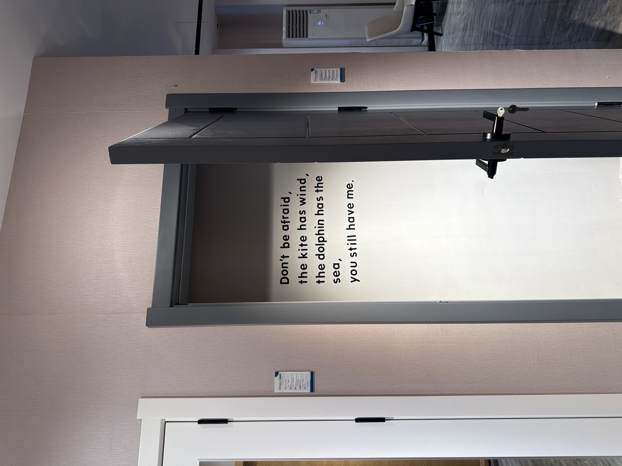 The image shows a partially open door with a message on it. The door is dark gray and has a doorknob with a key in it. The message on the door reads, "Don't be afraid, the kite has wind, the dolphin has the sea, you still have me." The door is set into a wall that has a beige, textured surface. Below the dark gray door is a white door. There are two small information labels on the wall, one above the gray door and one between the two doors. The door appears to be in a showroom or exhibition space, as the background includes glimpses of other exhibits, including an air conditioner unit and a white chair. The location is likely in Chancheng District, China, based on the text "gorge" being recognized which is one of the texts from the information labels in the image.
