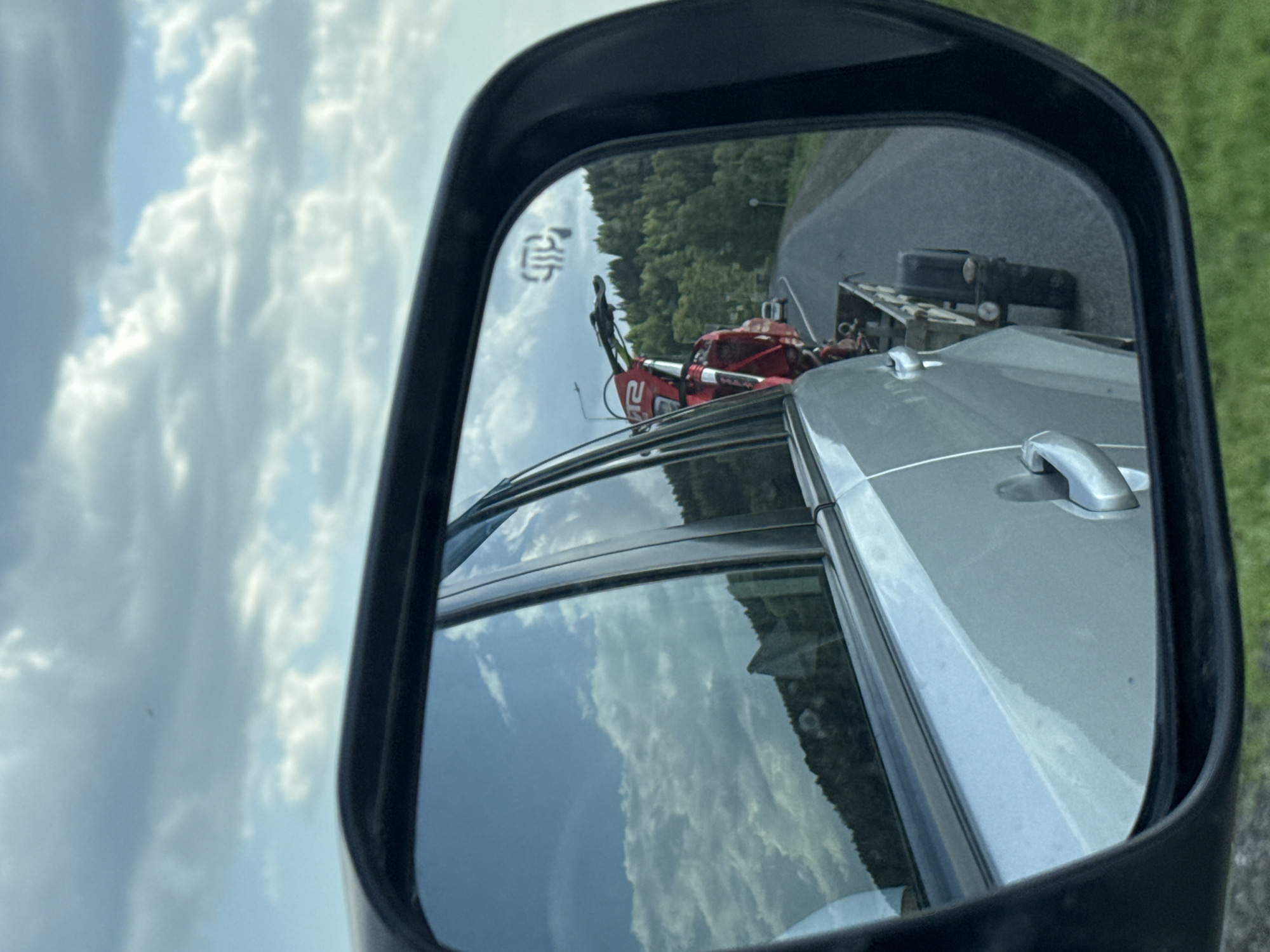 The image shows a side mirror of a vehicle reflecting a red dirt bike on a trailer being towed. The trailer has a black box-like object on it. The road is asphalt, with greenery visible in the background. The sky is visible as well with white puffy clouds. The vehicle door is also visible within the mirror's view. There is no evidence to suggest the image's location is in Ir'yak, Russia.