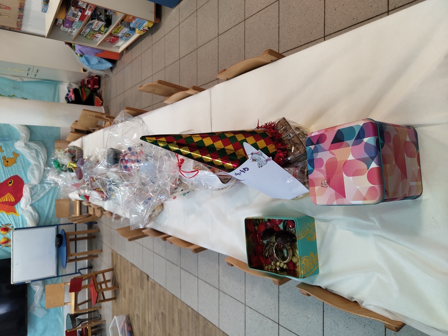 An indoor scene, likely a classroom or community center, is set up for a celebration. A long table, draped in a white tablecloth, stretches across the frame, laden with numerous wrapped gifts and decorative items. Wooden chairs line both sides of the table.
Many of the gifts are individually wrapped in clear cellophane, hinting at their contents. Prominently displayed near the center is a colorful party hat featuring a harlequin pattern in red, green, and gold, adorned with red tinsel fringe. Attached to the hat is a white paper tag with a small drawing of an animal and handwritten text, possibly "44" or "4:4". Adjacent to the hat, a large, rectangular tin box showcases a vibrant geometric pattern in shades of pink, blue, and white. Further down the table, a smaller, open teal-colored tin with gold accents reveals various items of jewelry, including necklaces, bracelets, and sunglasses.
The background to the left features a wall decorated with a blue fabric displaying white wave patterns and red and yellow fish, suggesting an aquatic or marine theme. Below this, smaller wooden tables and chairs are visible. In the upper left background, shelves stocked with various items, possibly toys or books, can be seen. The floor is covered with light-colored tiles.
The arrangement of party hats and numerous gifts indicates a festive event, likely a party or gathering, possibly for children, given the size of some furniture. The gifts appear to be party favors or prizes. The room is brightly lit, creating an inviting and celebratory atmosphere.