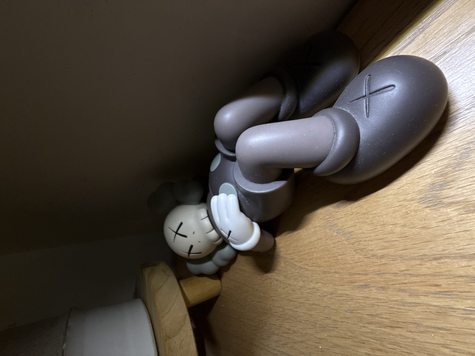 An indoor scene features a KAWS Companion vinyl art toy lying on a light brown wooden surface. The figurine is primarily dark brown with a light beige head, arms, and hands. Its head, featuring prominent "X" marks for eyes, is tilted slightly to the left. The figurine's body is posed in a supine position, with its left arm bent, hand resting near its chest, and its right arm extended to the side. Both legs are bent at the knees. A distinctive "X" mark is visible on the bottom of the right foot. To the upper left, a portion of a white cylindrical object rests on a small, light wooden stand with short, visible legs. The background and the right side of the image consist of a plain, dark brown vertical surface, suggesting a wall or the interior of a shelf. The scene is dimly lit, creating soft shadows. The specific location within the city of New York, United States of America, cannot be determined from the visual information provided.