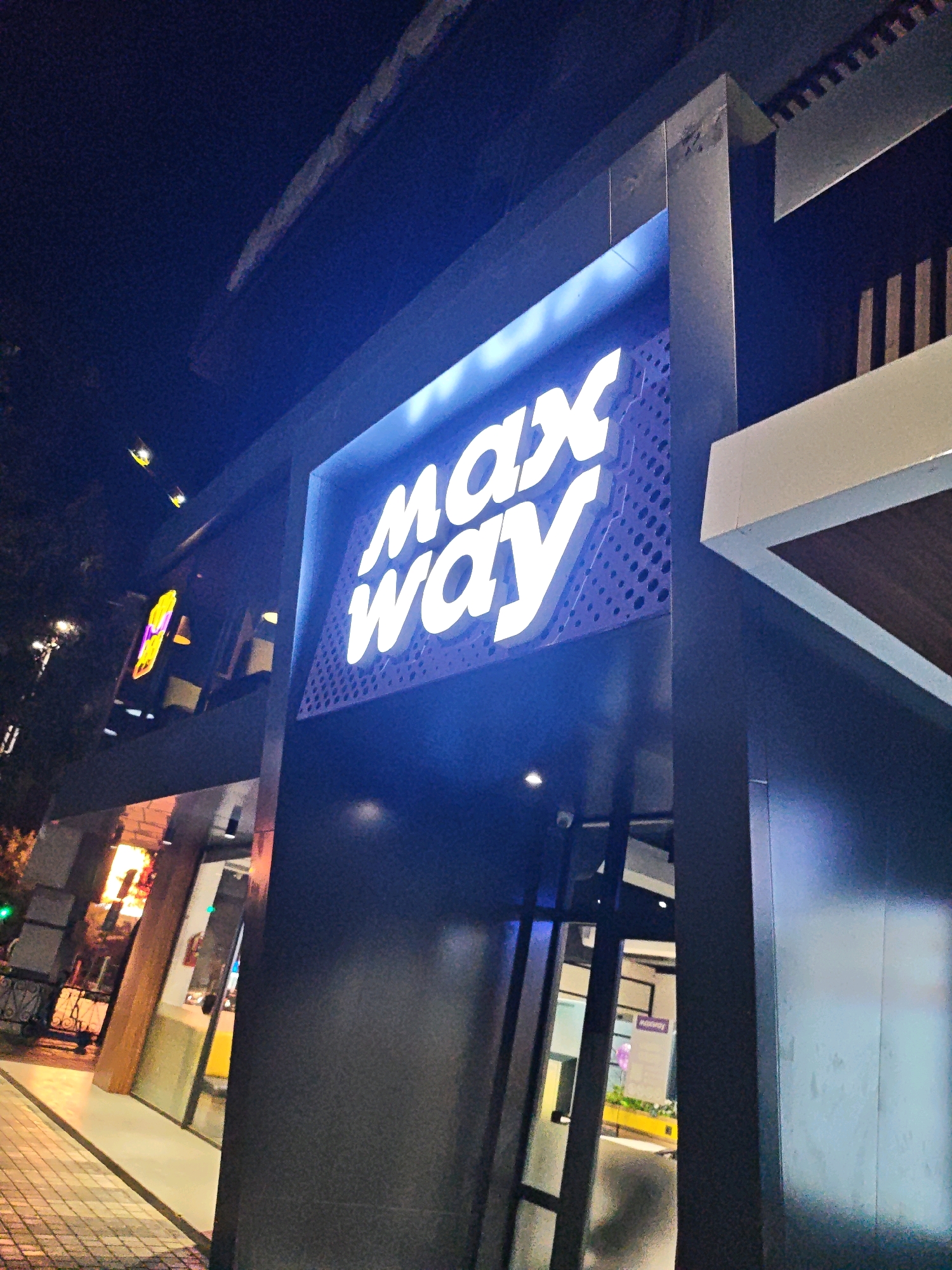 A nighttime exterior shot captures the brightly lit "max way" sign of a business establishment. The sign, in white lettering with a blue dotted background, is prominently displayed on the dark facade of the building. The architecture appears modern, with a dark gray, angular exterior. To the left, a glimpse of the interior reveals yellow and white elements, possibly seating or displays. The street in front has a tiled sidewalk illuminated by streetlights. In the background, trees and other illuminated signs suggest a busy urban environment. The image is likely taken in Tashkent, Uzbekistan, as indicated by a previous analysis.