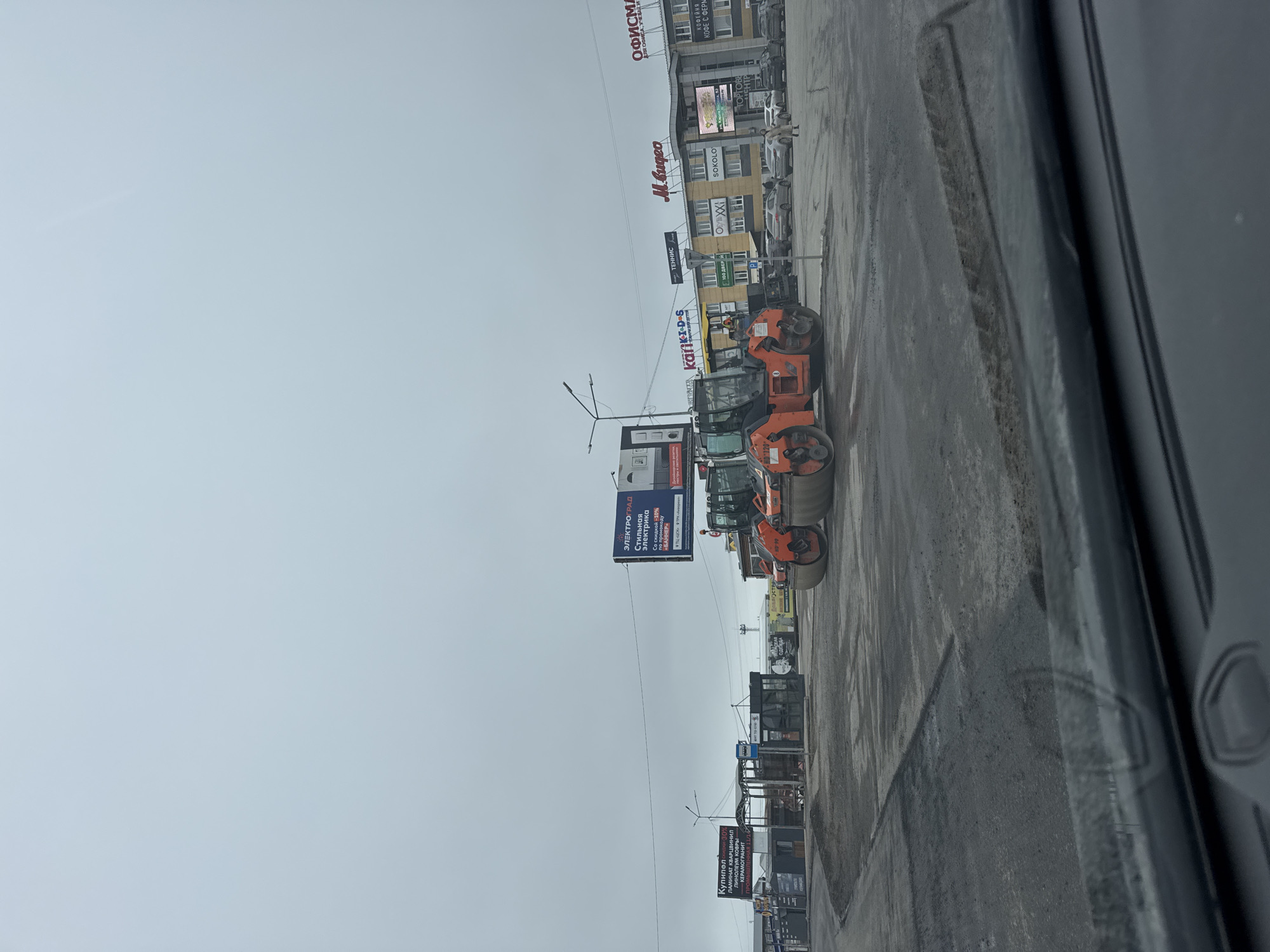 The image, taken from inside a vehicle, shows a street in Chaikovsky, Russia. The sky is overcast. Along the right side of the street are several orange road rollers. Billboards and store signs are visible on the left side of the street, including signs for "Kupinel," "Elektrograd," "Kariki DS," "Tennis," "M.Video," "Sokolo," and "Ofisma." The road appears wet and recently paved.