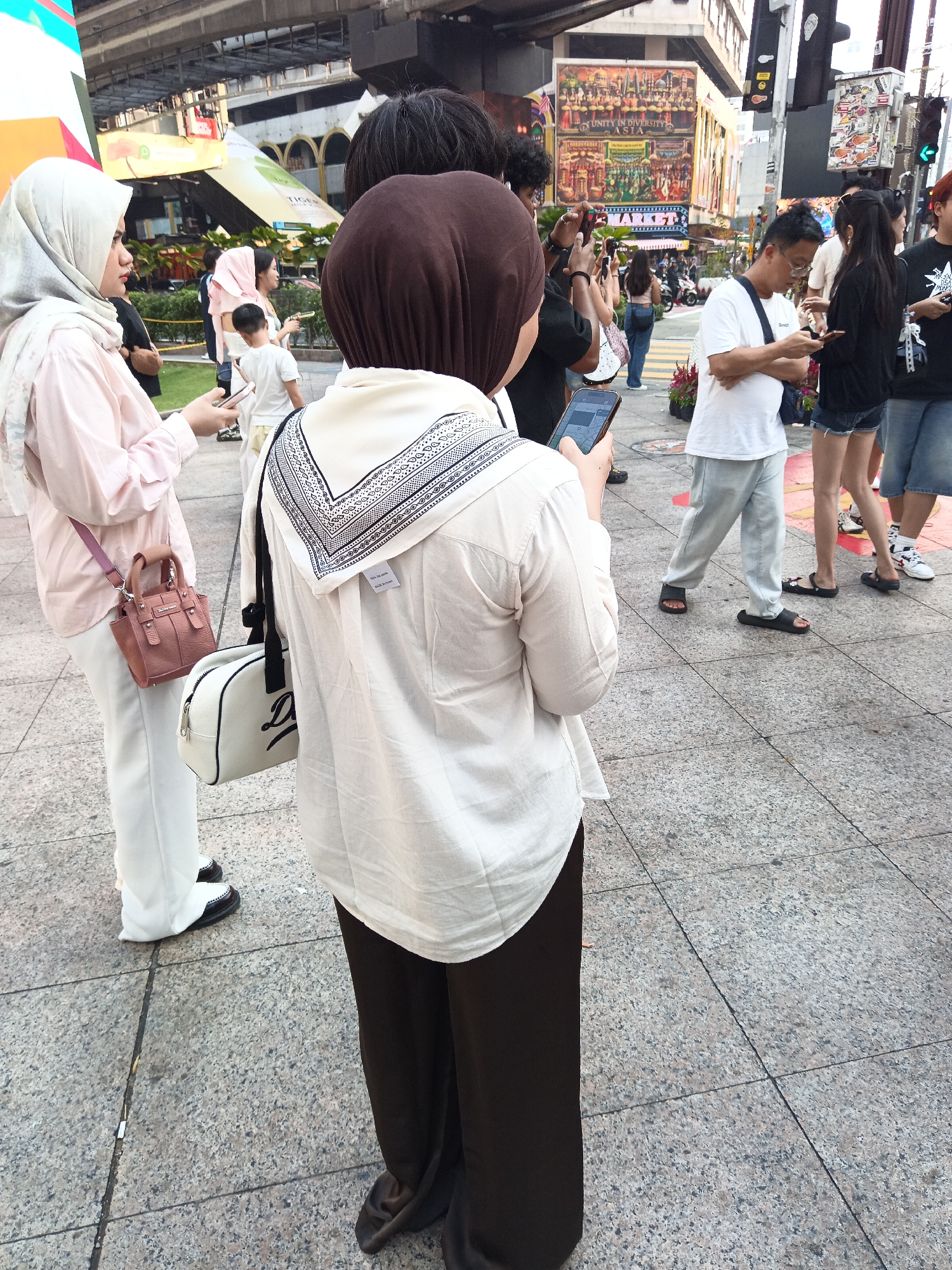 This is a medium shot of a crowd of people on a sidewalk in Kuala Lumpur, Malaysia. The scene appears to be near a busy street with buildings and billboards in the background. In the foreground, a woman wearing a hijab and a light pink long-sleeved shirt is holding a pink handbag and a smartphone. To her right, another woman, facing away from the camera, wears a brown hijab and a white long-sleeved shirt with a patterned scarf around her neck. She is also holding a smartphone. A white bag with black lettering is slung over her shoulder. Other people are visible in the background, some walking and others standing and looking at their phones. One man in a white t-shirt and light gray pants is looking down at his phone. The sidewalk is paved with large, speckled gray tiles. The overall impression is of a bustling urban environment.