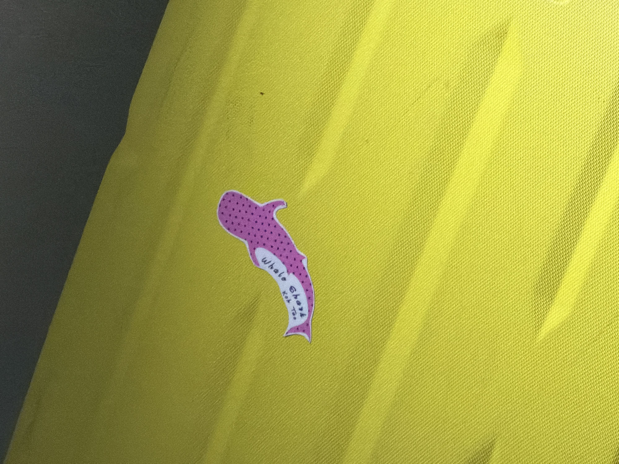 A sticker of a pink whale shark with purple polka dots is placed on a bright yellow, textured surface, possibly fabric. The whale shark sticker has the words "Whale Shark Kid Too" written on it in a black handwritten font. The background is split between the yellow surface and a dark gray area, suggesting a shadow or different material. The image appears to be a close-up, focusing on the sticker and the yellow surface it rests on. There is no discernible context or information about its location in Baan Mae Nam, Thailand within the visual data.