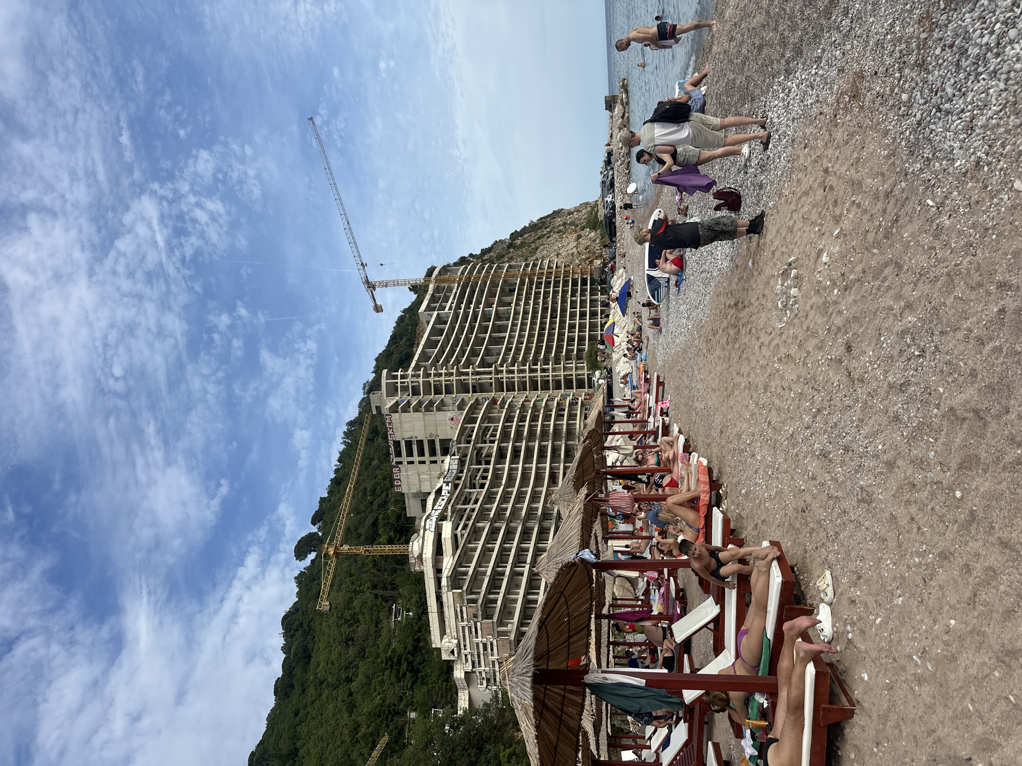 This image captures a vibrant beach scene in Mirište, Montenegro, on what appears to be a sunny day with scattered white clouds across a bright blue sky. The composition is taken from a slightly elevated perspective, looking towards the sea and a hillside.
In the foreground and midground, a pebble and sand beach is bustling with activity. Numerous beachgoers are visible, with many relaxing on sun loungers arranged in rows, some under thatched umbrellas that provide shade. People are seen sunbathing, sitting, and walking on the beach. Towards the water's edge, several individuals are in the clear blue sea, either standing or swimming. Two men are standing on the shoreline, one wearing a black t-shirt and camouflage shorts, and another in a white t-shirt and grey shorts with a black backpack.
A significant construction project dominates the left side of the image. A large, multi-story building, still in its concrete skeleton phase, is under construction on the side of a green, tree-covered hill. Two tall yellow construction cranes are prominently featured, towering over the building and reaching into the sky, indicating ongoing development. One of the cranes is positioned near the peak of the hill, while the other is closer to the building.
Visible text on the construction site includes "EDER" and what appears to be "MESRER" or a similar word, possibly graffiti or company names. The presence of tourists and the construction suggests a developing resort area. The overall mood is relaxed and recreational, despite the imposing construction in the background.
