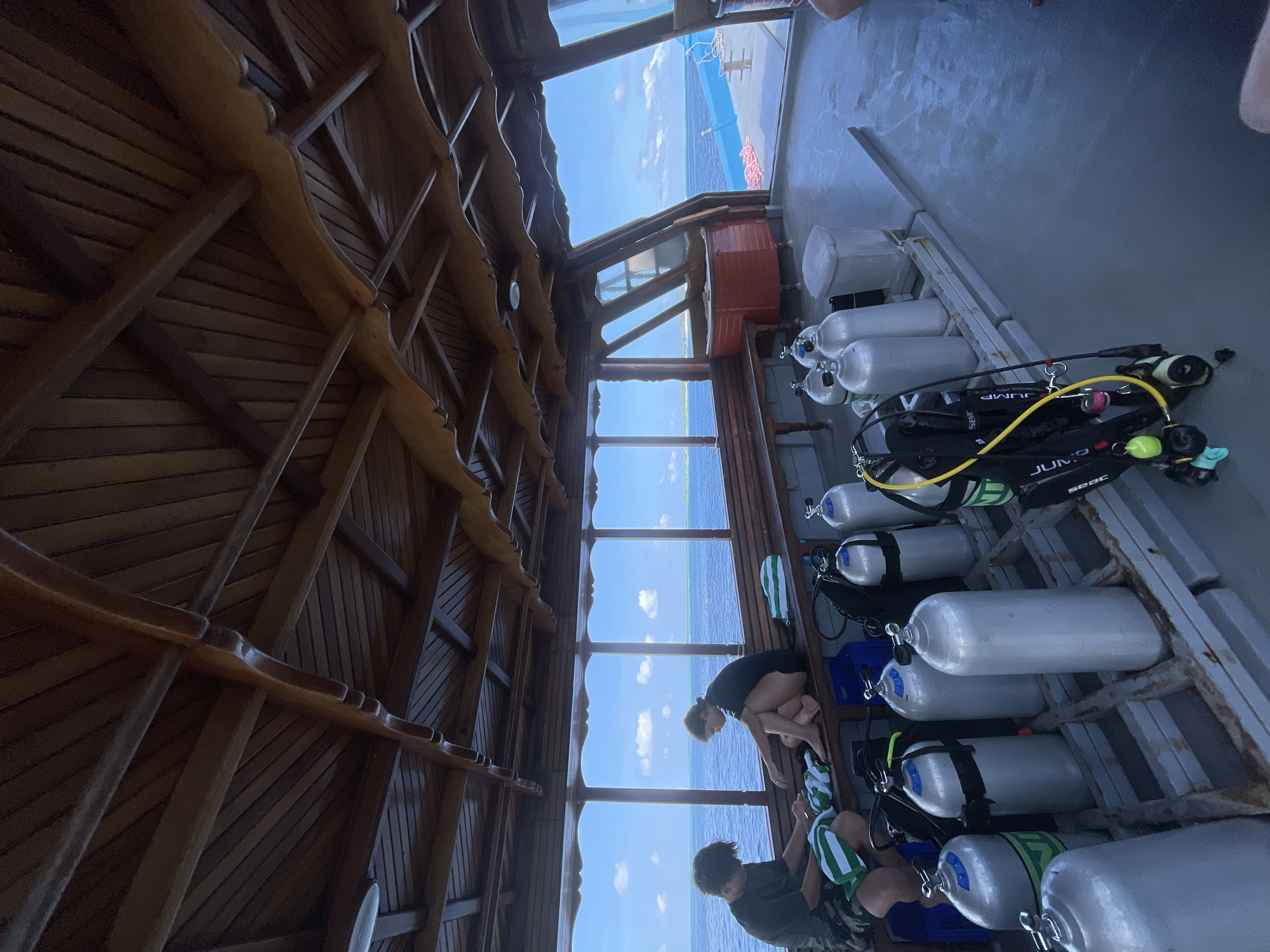 On a wooden boat deck, two individuals are seated on a wooden bench facing the ocean. The individual on the left, wearing a dark t-shirt and patterned shorts, is looking down at a striped green and white towel. The individual on the right, wearing a black t-shirt and shorts, is also looking down. Several silver scuba tanks are neatly arranged on the deck in the foreground and midground, with some black dive gear and a blue crate also visible. The boat is on the water, with the ocean and a distant island visible through wooden-framed windows. The ceiling of the boat is constructed from dark wood with visible beams. The environment appears to be a sunny day with a clear blue sky and scattered clouds.
