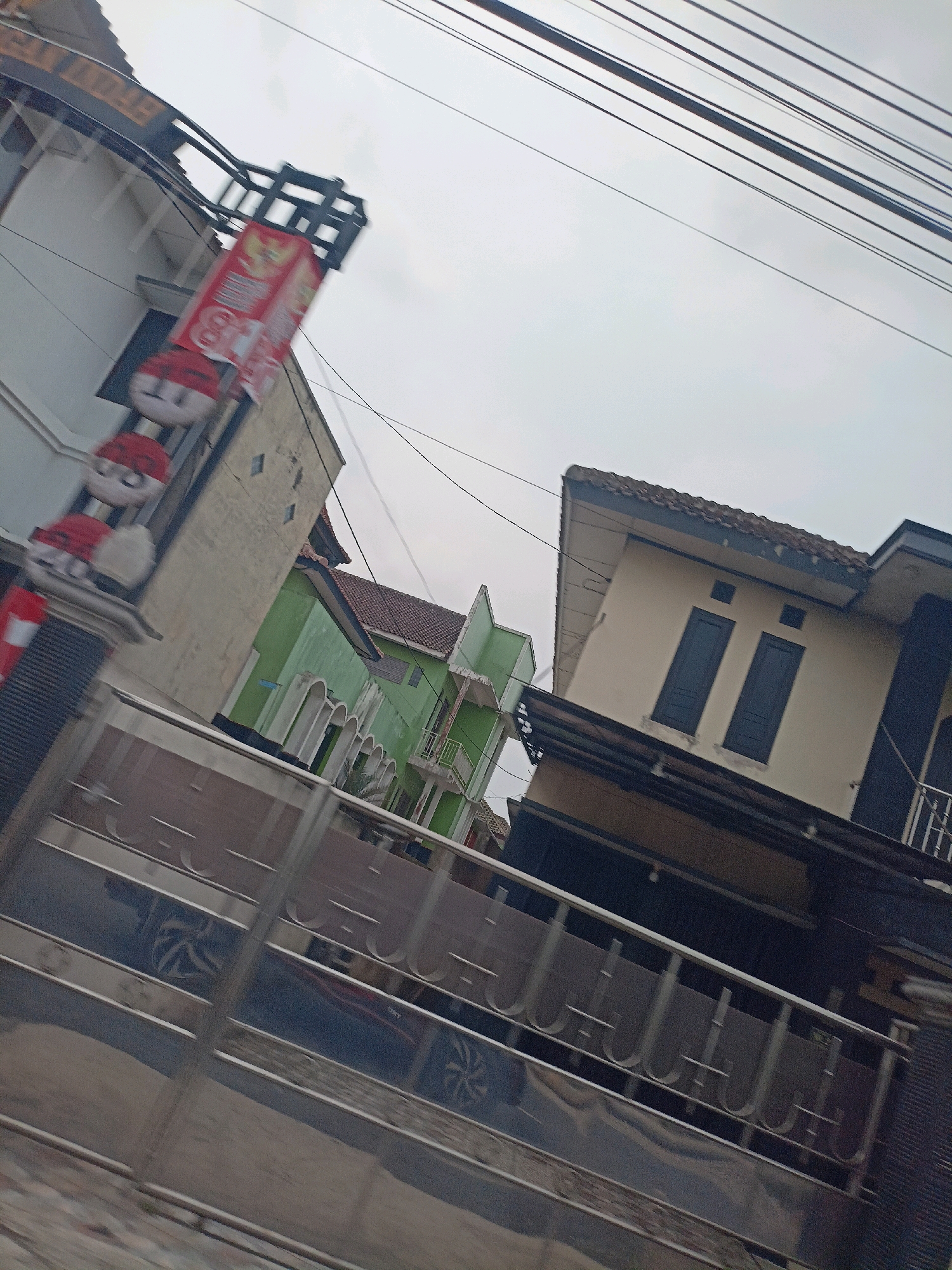 The image shows a street scene in Bandongan, Indonesia, with several buildings and a metal fence in the foreground. The buildings appear to be houses or small commercial establishments. One building on the left has a sign with red and white decorations, possibly related to a local event or business. Another building in the middle is painted green with arched openings. A beige building with dark windows is visible on the right. The sky is overcast. The street is paved, and there are power lines overhead. The metal fence in the foreground has a decorative pattern.