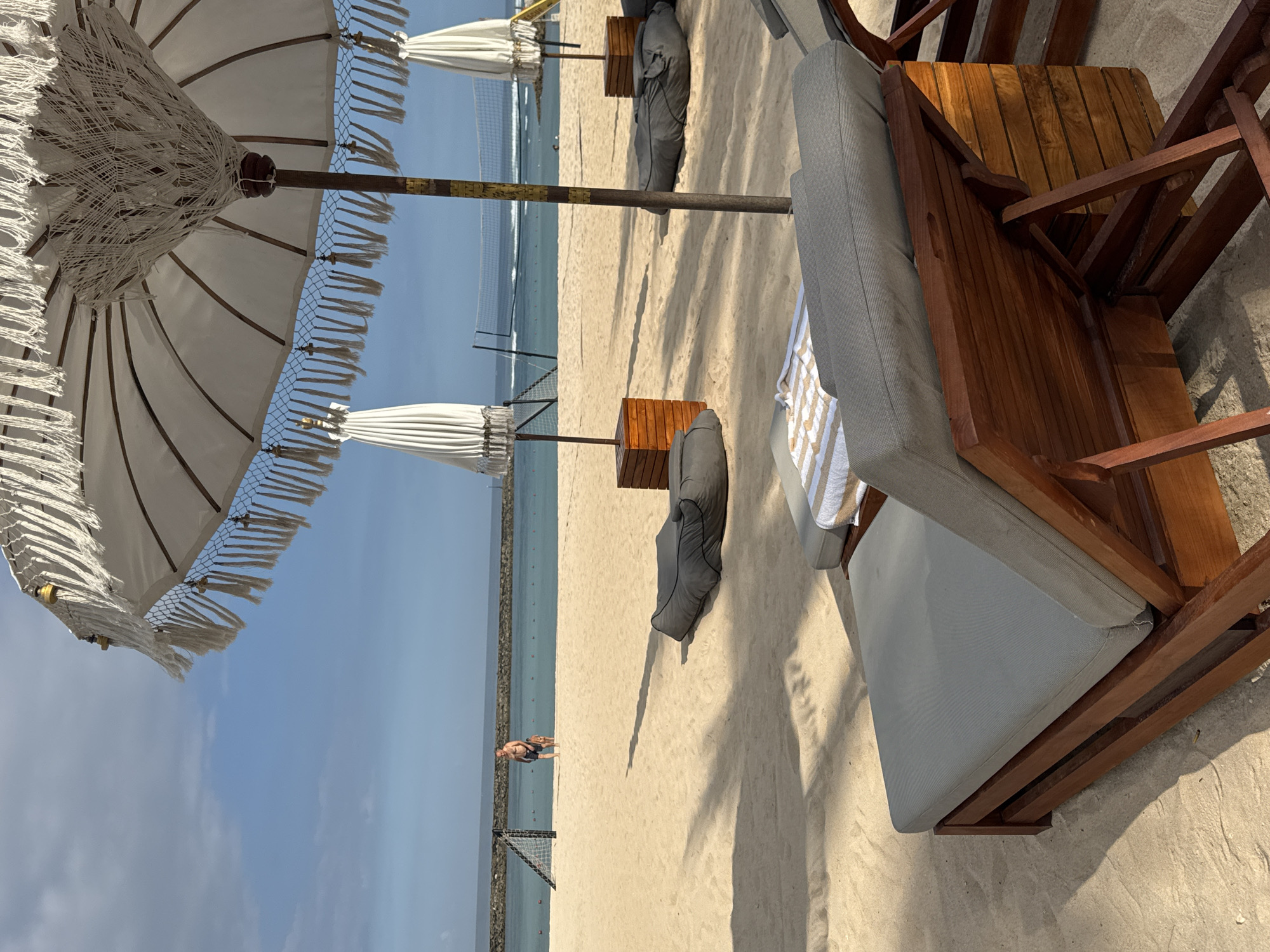 This image depicts a beach scene in Kuta Selatan, Indonesia. Visible are sandy shores, the ocean, and a section of the sky. Beach furniture includes lounge chairs with gray cushions, wooden side tables, and large white umbrellas with fringed edges. Further down the beach, there are additional umbrellas and beanbag chairs. A man is walking along the water's edge. The scene is lit by sunlight, casting shadows from the umbrellas and furniture onto the sand. A net is present at the far end of the beach.