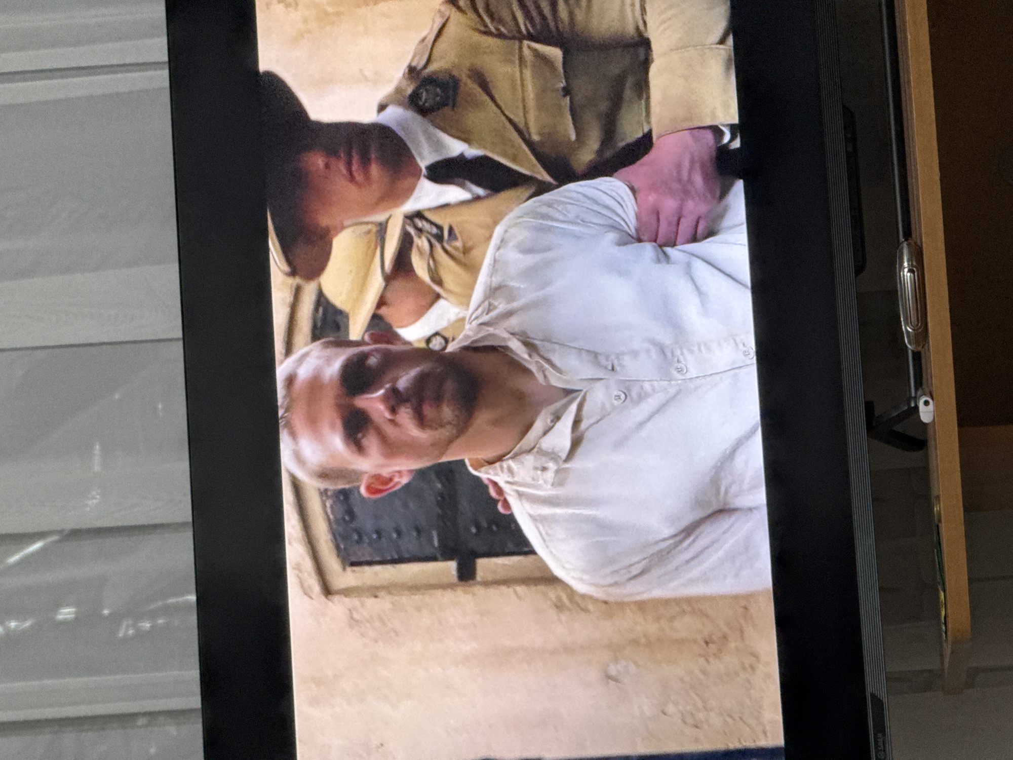 The image shows a close-up of a scene from a movie or television show, with a man in a white shirt being held by another man in a khaki uniform. The man in white has short, light brown hair and a serious expression. The man in uniform, who is wearing a hat, has his arm around the man in white's shoulders. The background appears to be a stone wall. There is no visible indication that this scene is located in the city of Valuyki, Russia, or any other specific location.