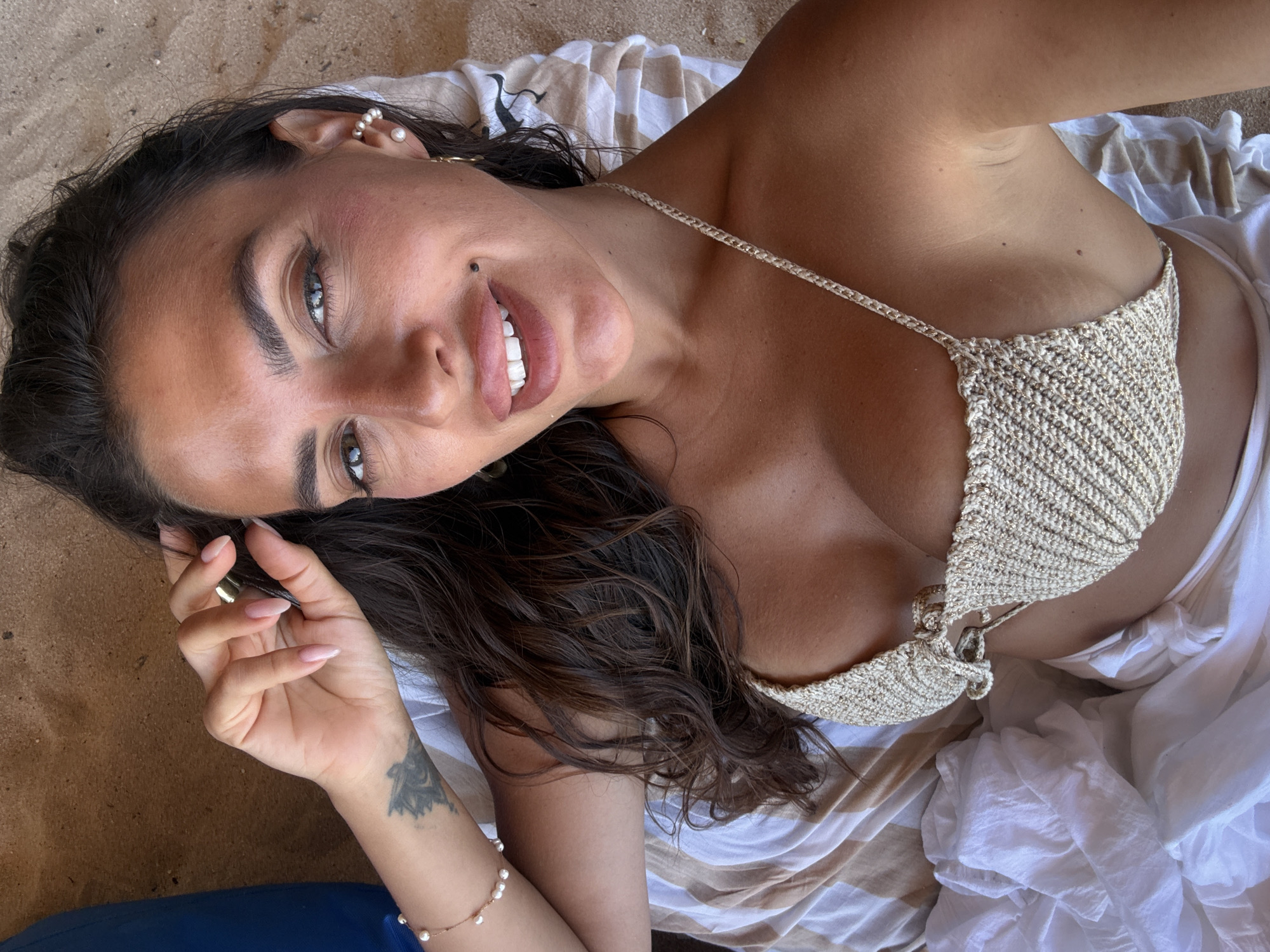 A close-up, overhead shot captures a woman lying on her back on a beach in Kuta Selatan, Indonesia. She has long, dark, wavy hair that is spread around her face. Her skin is tanned, and she is wearing a light-colored, crocheted bikini top with gold accents. She has full lips, prominent cheekbones, and dark eyebrows. A small mole is visible below her lip. Her left arm is bent, with her hand resting near her head, displaying a light pink manicure. A delicate pearl bracelet is on her left wrist, and a tattoo is visible on her forearm. She is wearing a pearl earring in her right ear. She is lying on a striped towel that is partially covered by white fabric. The background appears to be sand.